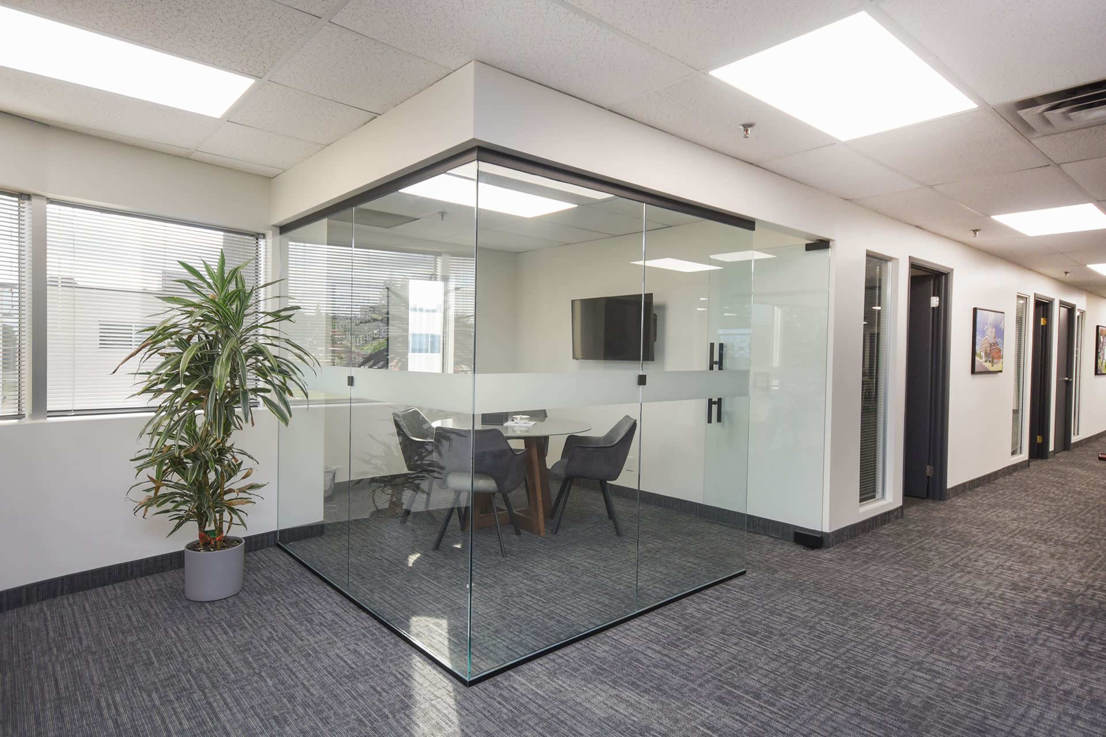 The image shows a modern office space featuring a glass-walled meeting room with a table and chairs, surrounded by a carpeted hallway with offices on either side.