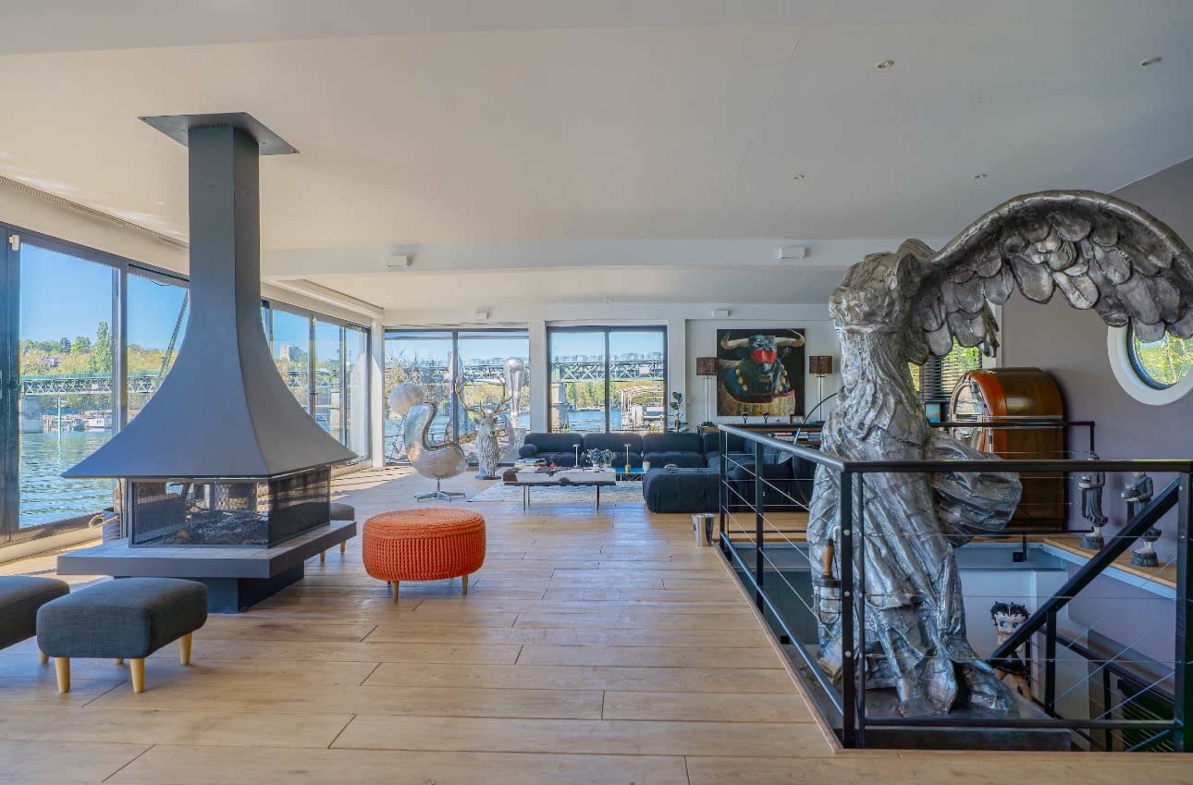 The image shows a modern, open-concept living space with large windows overlooking a body of water, featuring a prominent fireplace and an ornate sculpture of an angel.