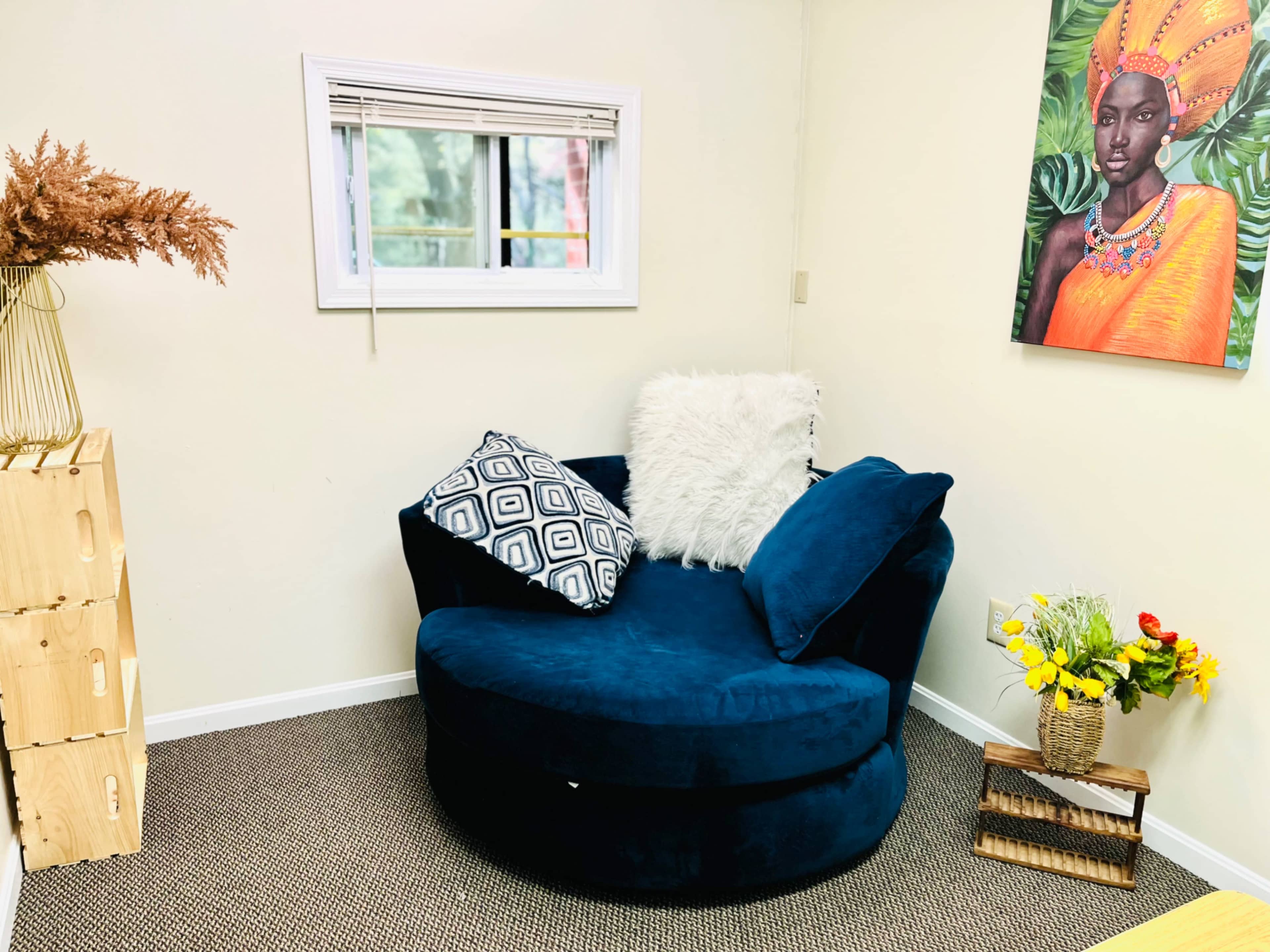 A cozy corner featuring a circular blue chair with a white fur pillow, a patterned black and white pillow, a small wooden shelf with flowers, and a framed portrait on the wall.