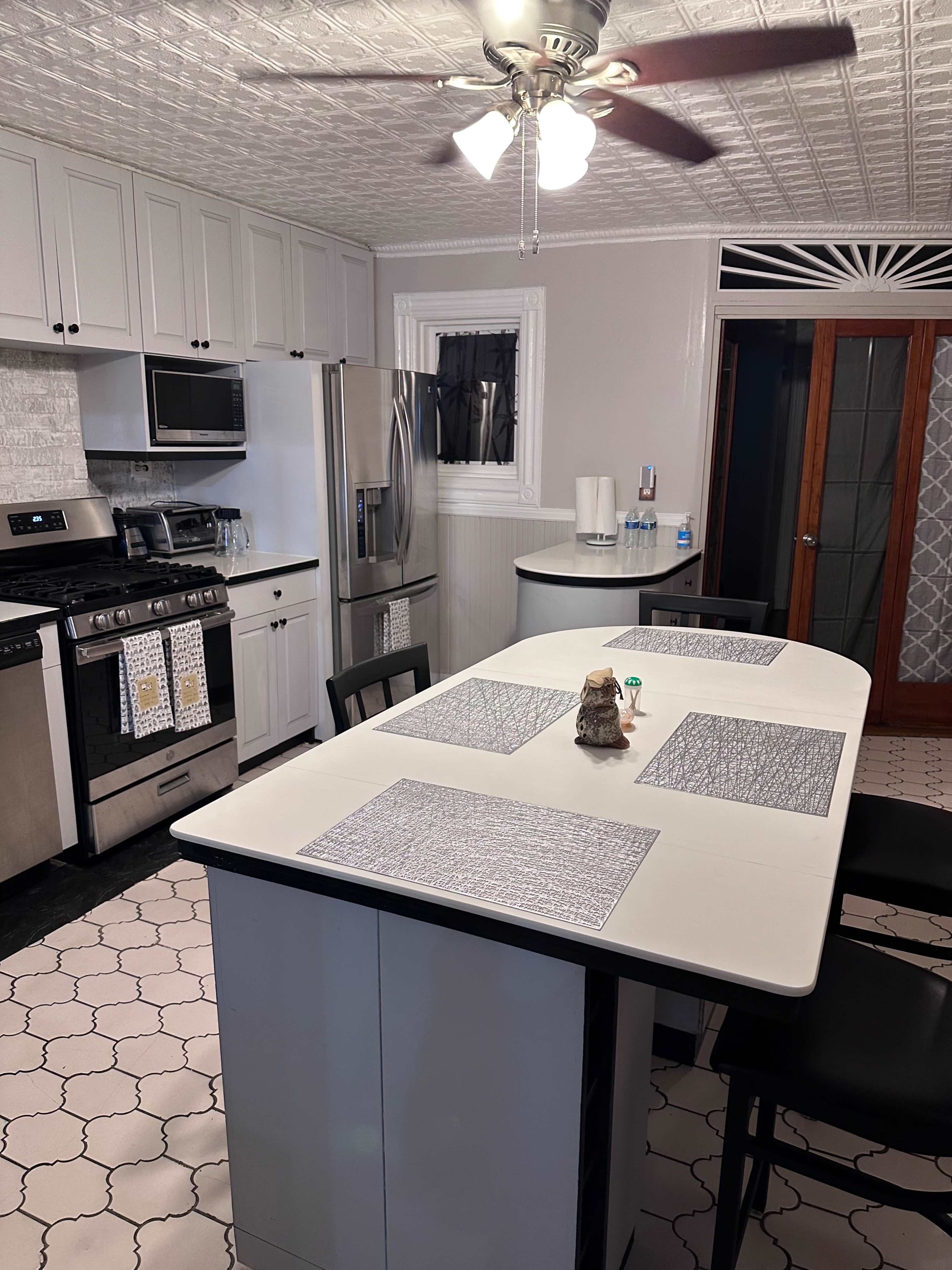 The image shows a modern kitchen with light-colored cabinets, stainless steel appliances, and a central island with black chairs and placemats.