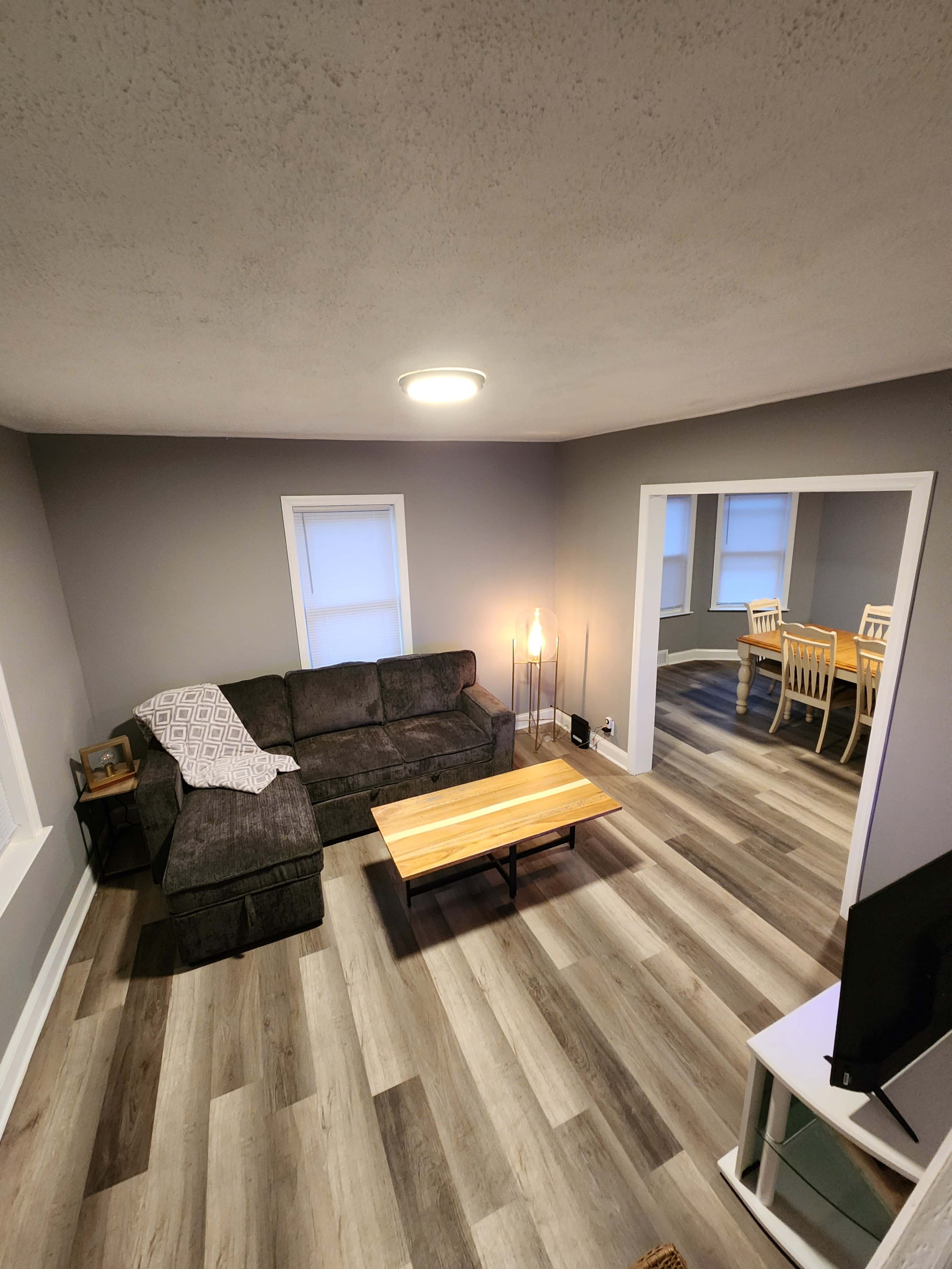 A cozy living room features a dark gray sectional sofa, a wooden coffee table, and a dining area with a table and chairs visible through an archway.