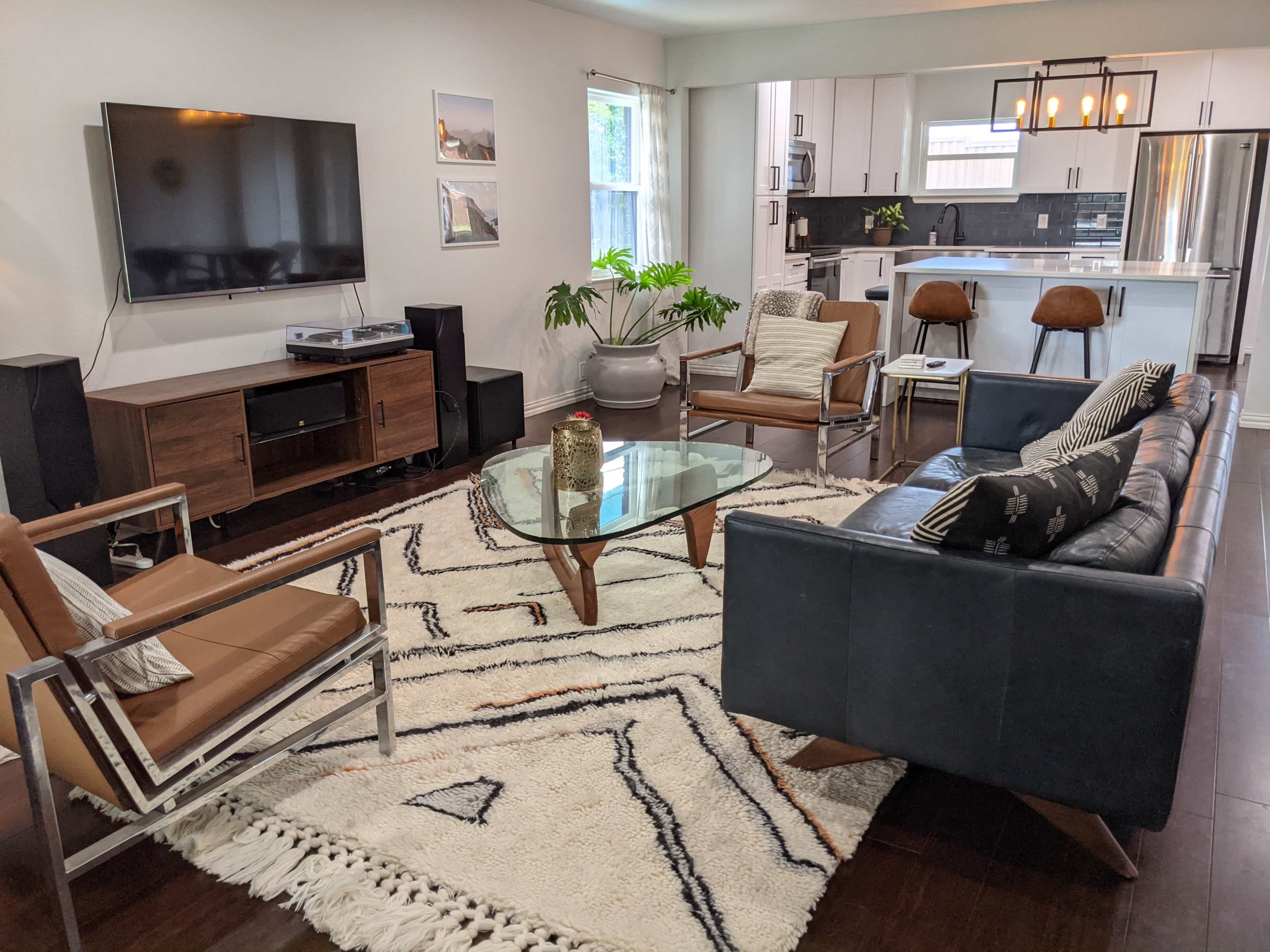 A modern living room with a glass coffee table, two armchairs, a sectional sofa, and a television mounted on the wall, alongside a kitchen area in the background.