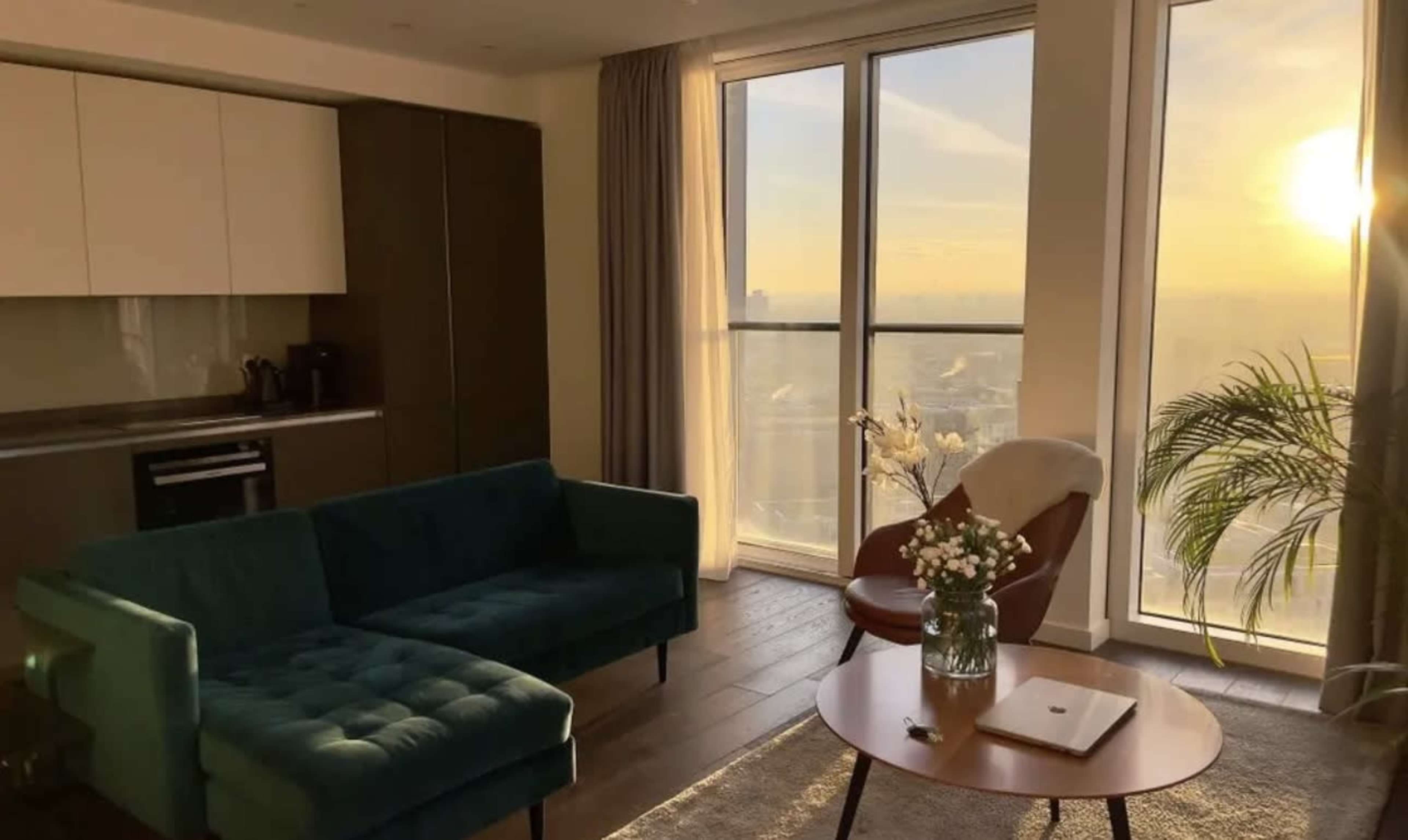 A modern living room features a teal sofa, a round wooden coffee table with a laptop, and large windows showcasing a sunset view.