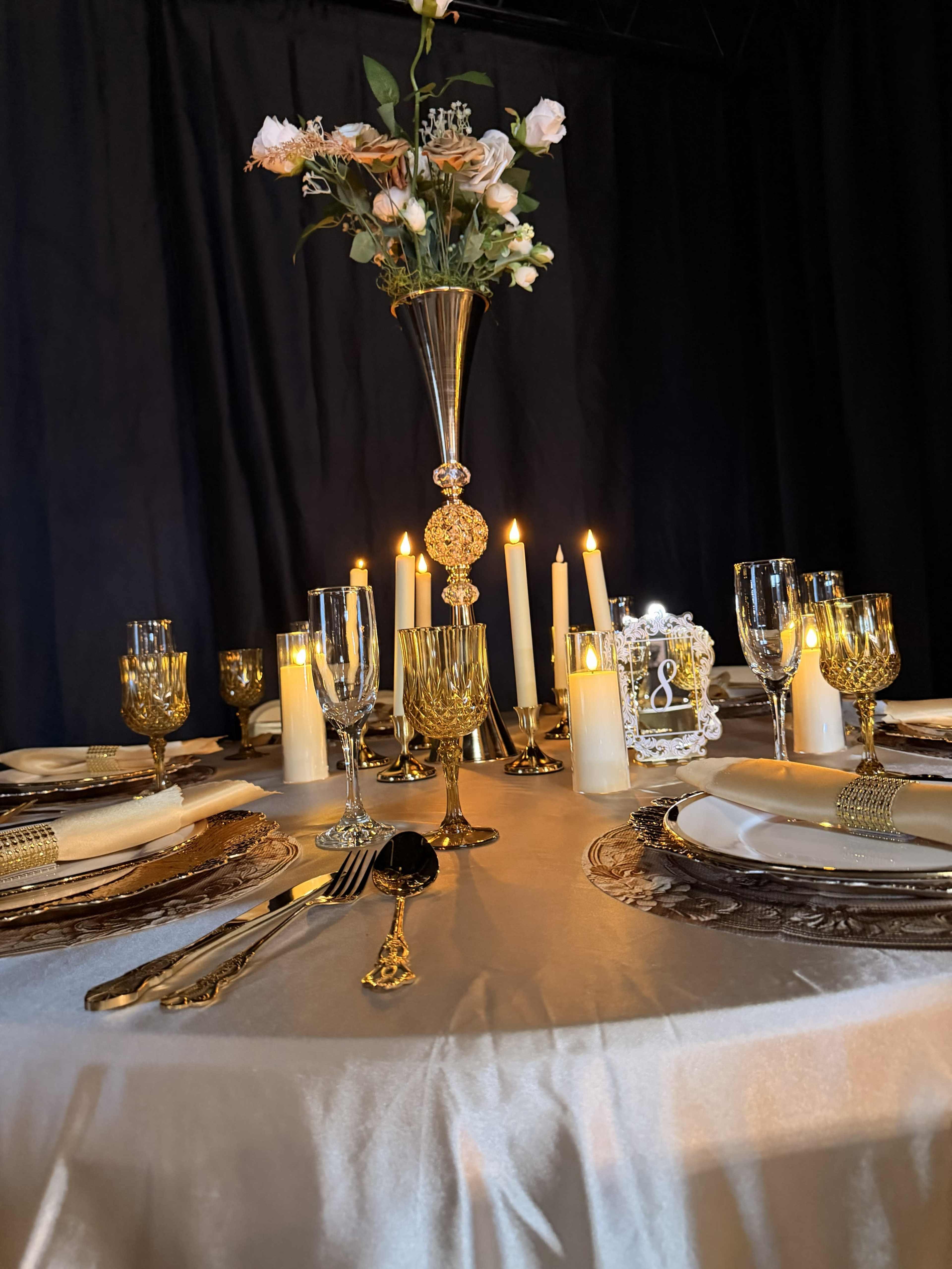 A round dining table is elegantly set with gold-accented tableware, a tall floral centerpiece, and candles illuminating the dark backdrop.