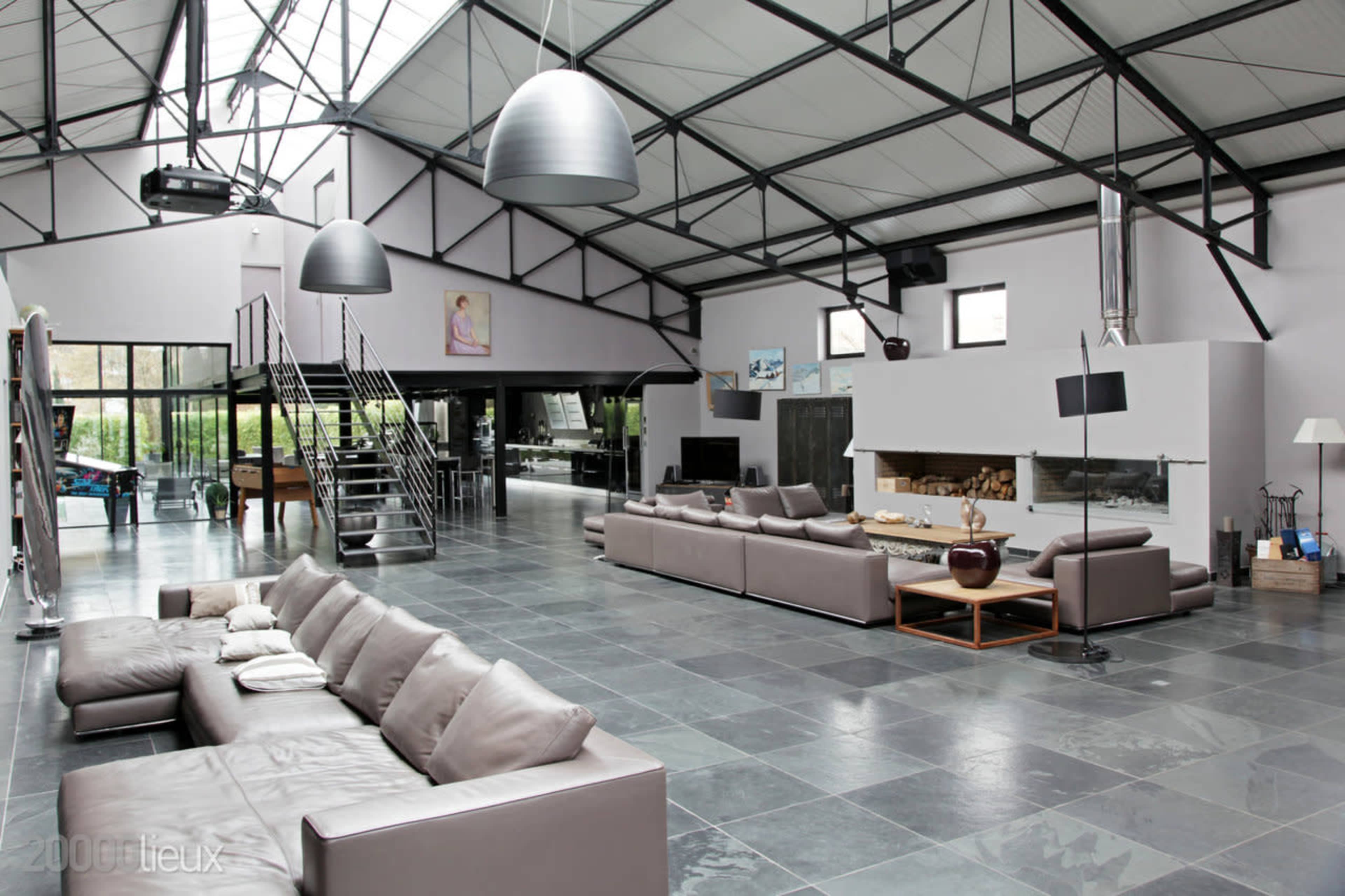 A spacious, modern loft interior featuring leather sofas, high ceilings with metal beams, and large windows allowing natural light.