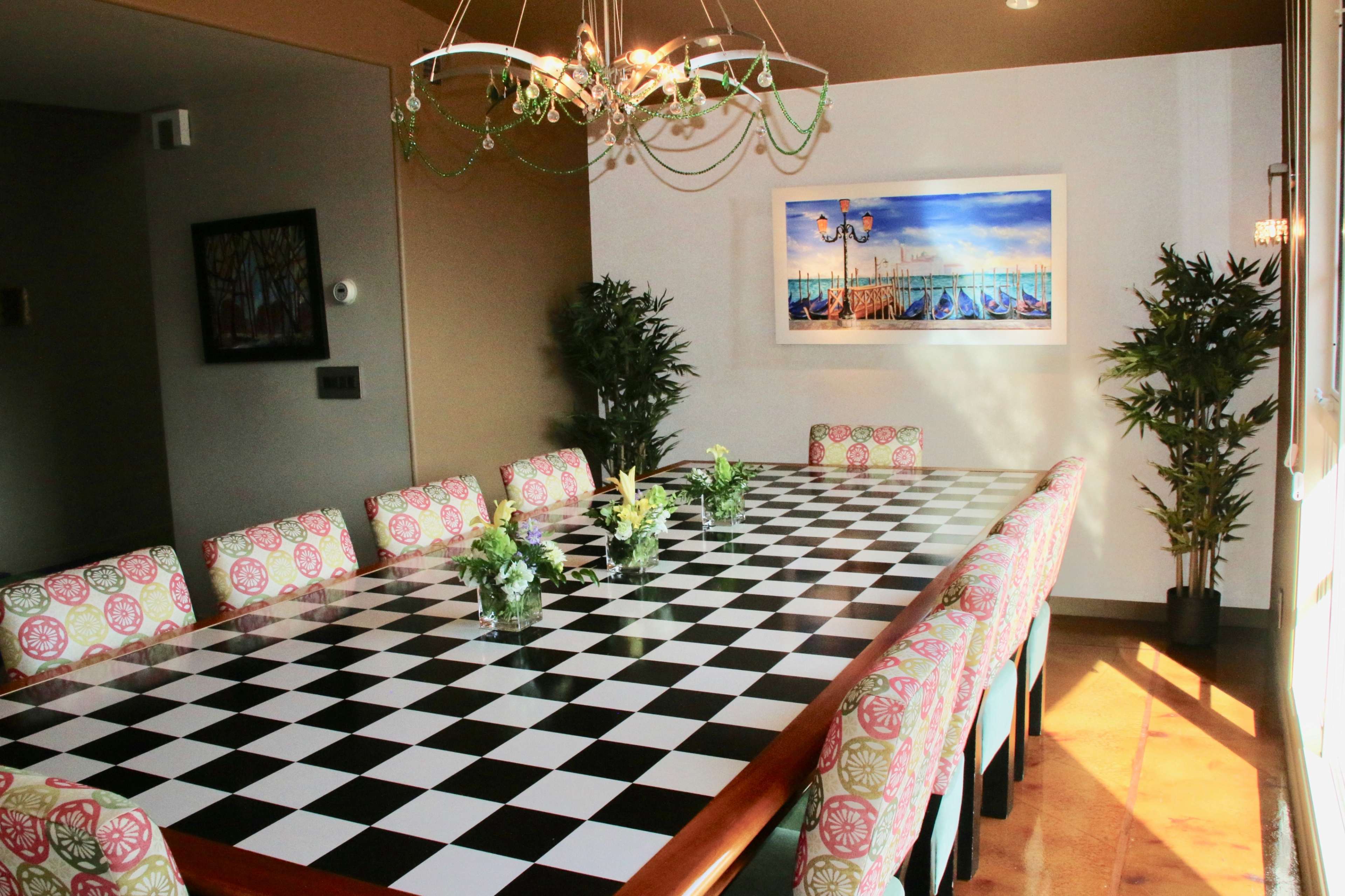 A large dining table with a black and white checkered top is surrounded by chairs featuring floral-patterned cushions, set in a brightly lit room with plants and artwork on the walls.