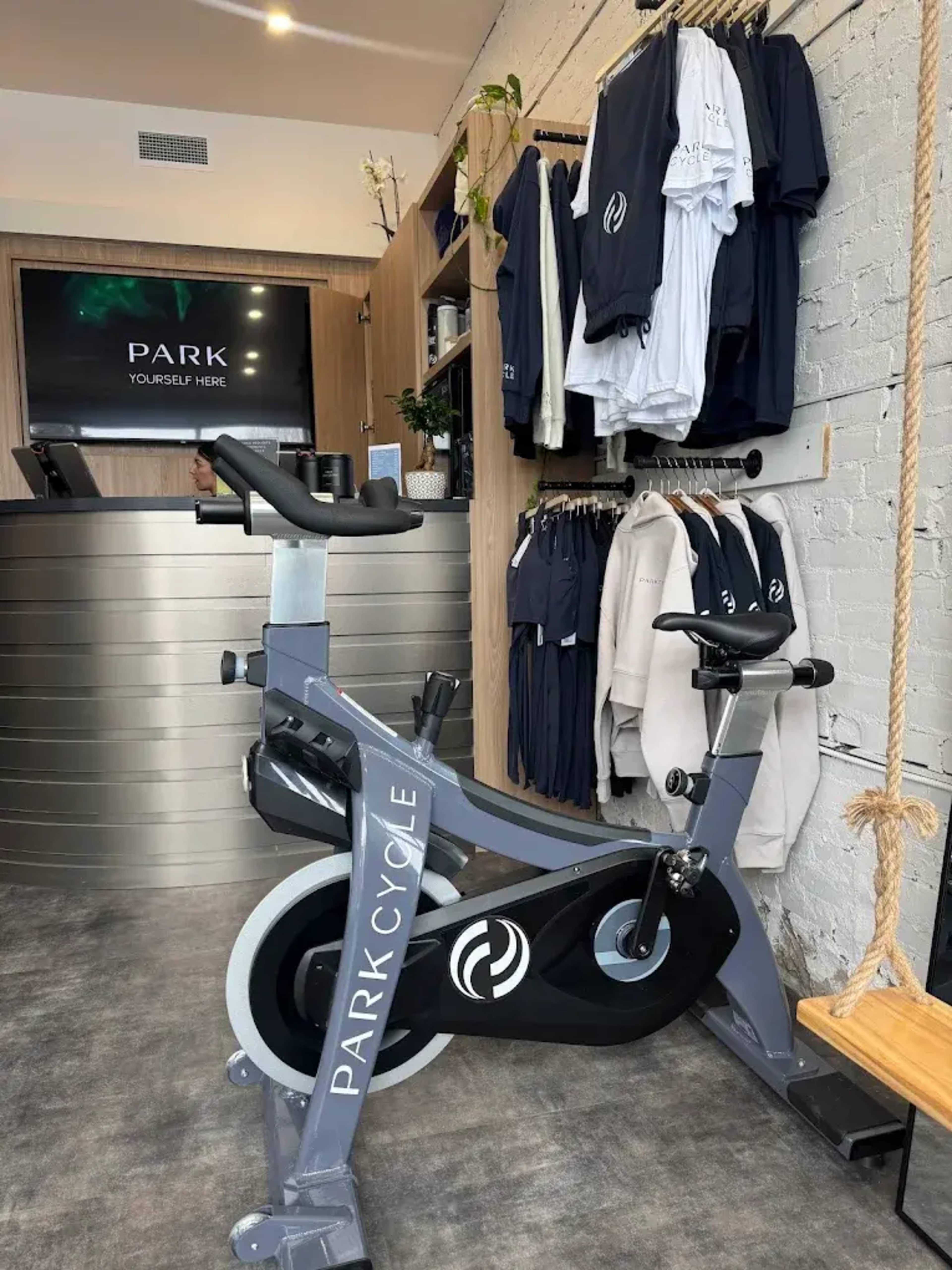 The image shows a stationary bike in a fitness studio, with merchandise displayed on the wall and a reception area in the background.