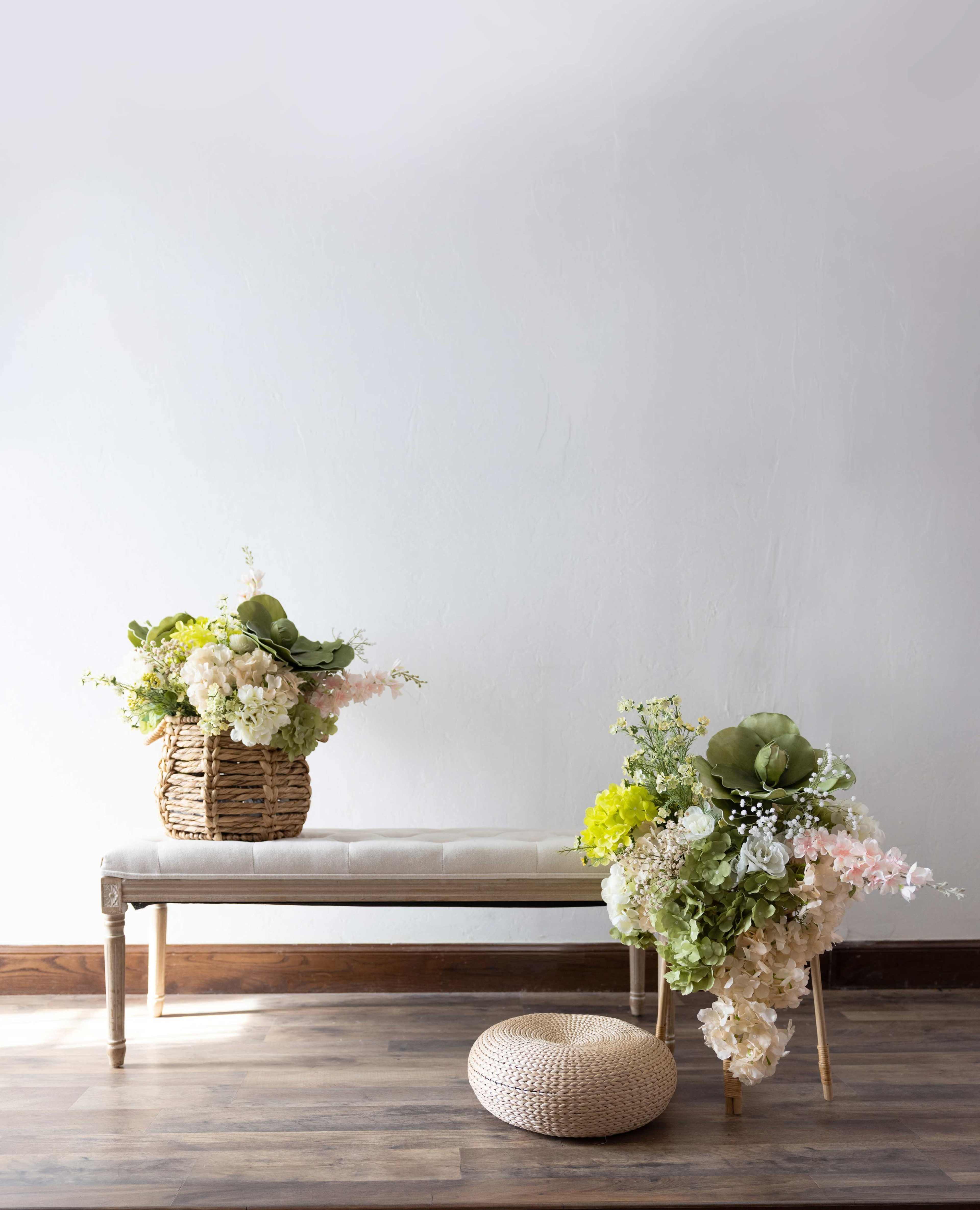 A bench with a soft cushion displays two large floral arrangements in woven baskets beside a round pouf on a wooden floor.