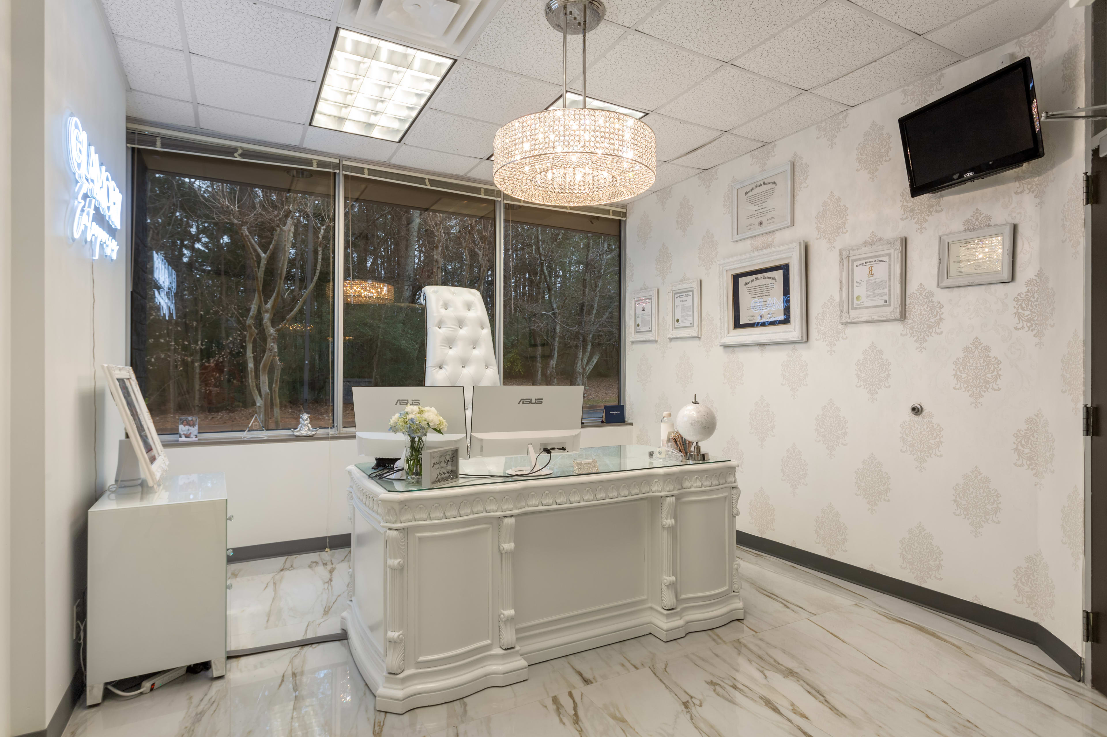 A modern office interior with a white desk, a high-backed chair, and a decorative chandelier, surrounded by framed certificates and a large window.