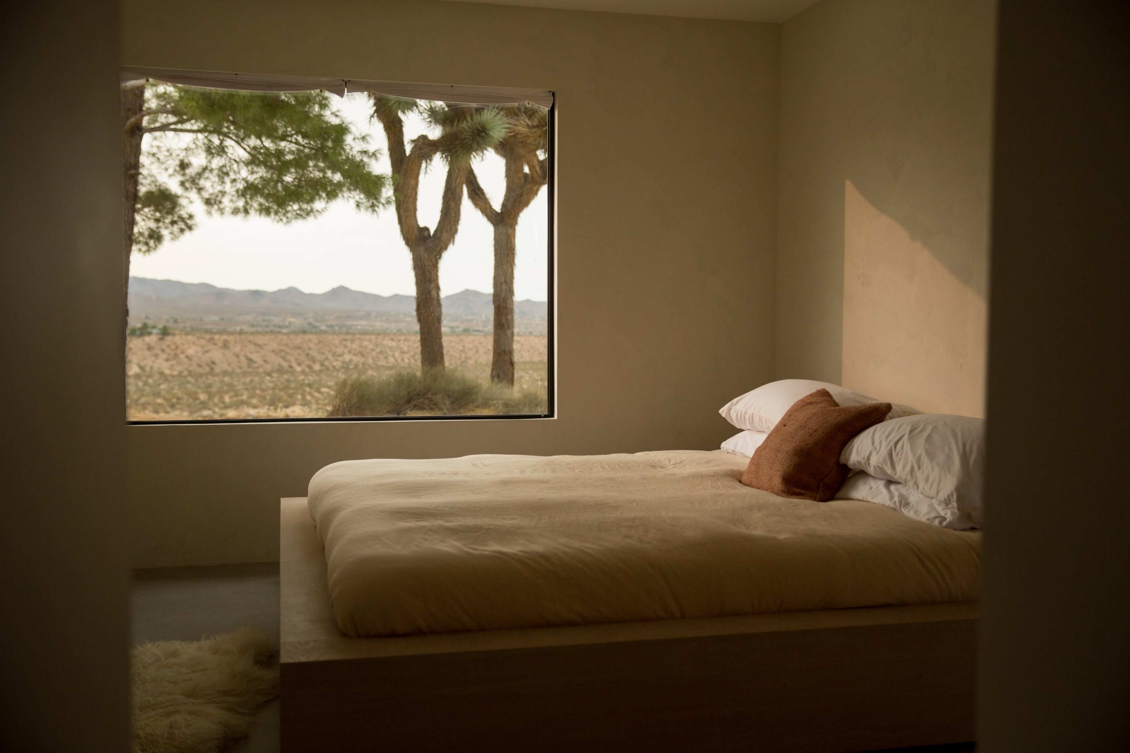 A minimalist bedroom features a simple bed and a large window showcasing a view of trees and an arid landscape.