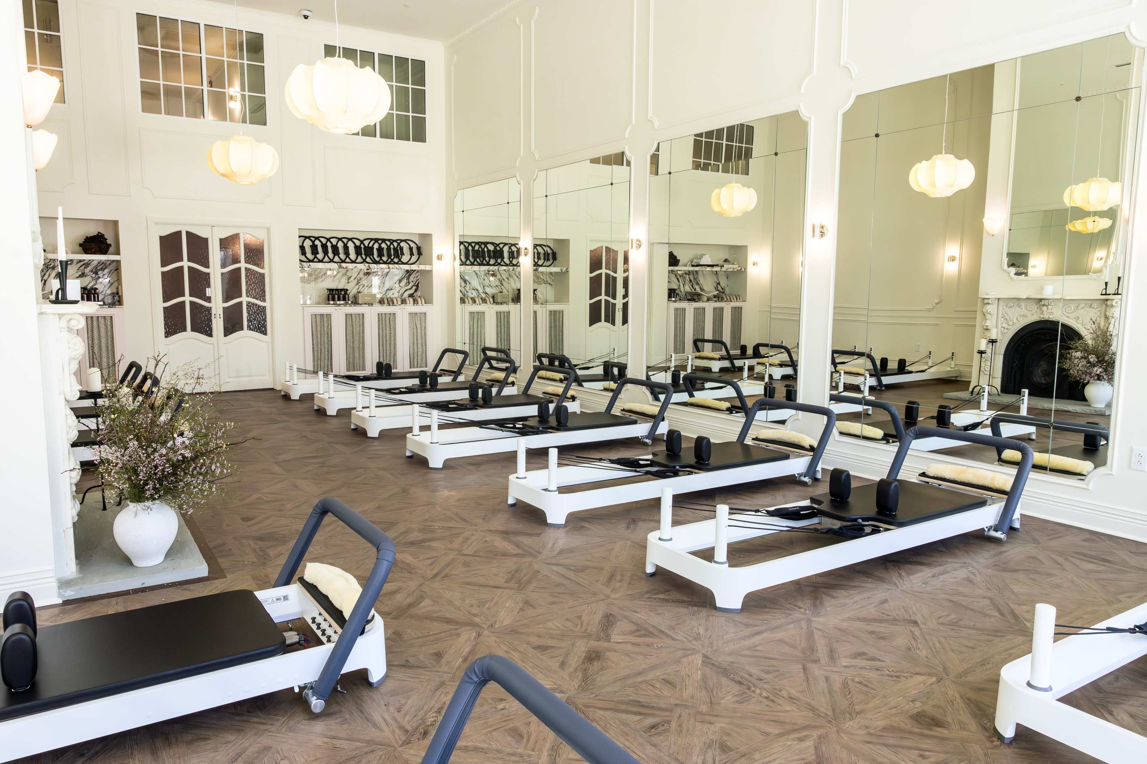The image shows a bright, spacious pilates studio with reformer machines arranged neatly in a mirrored room.