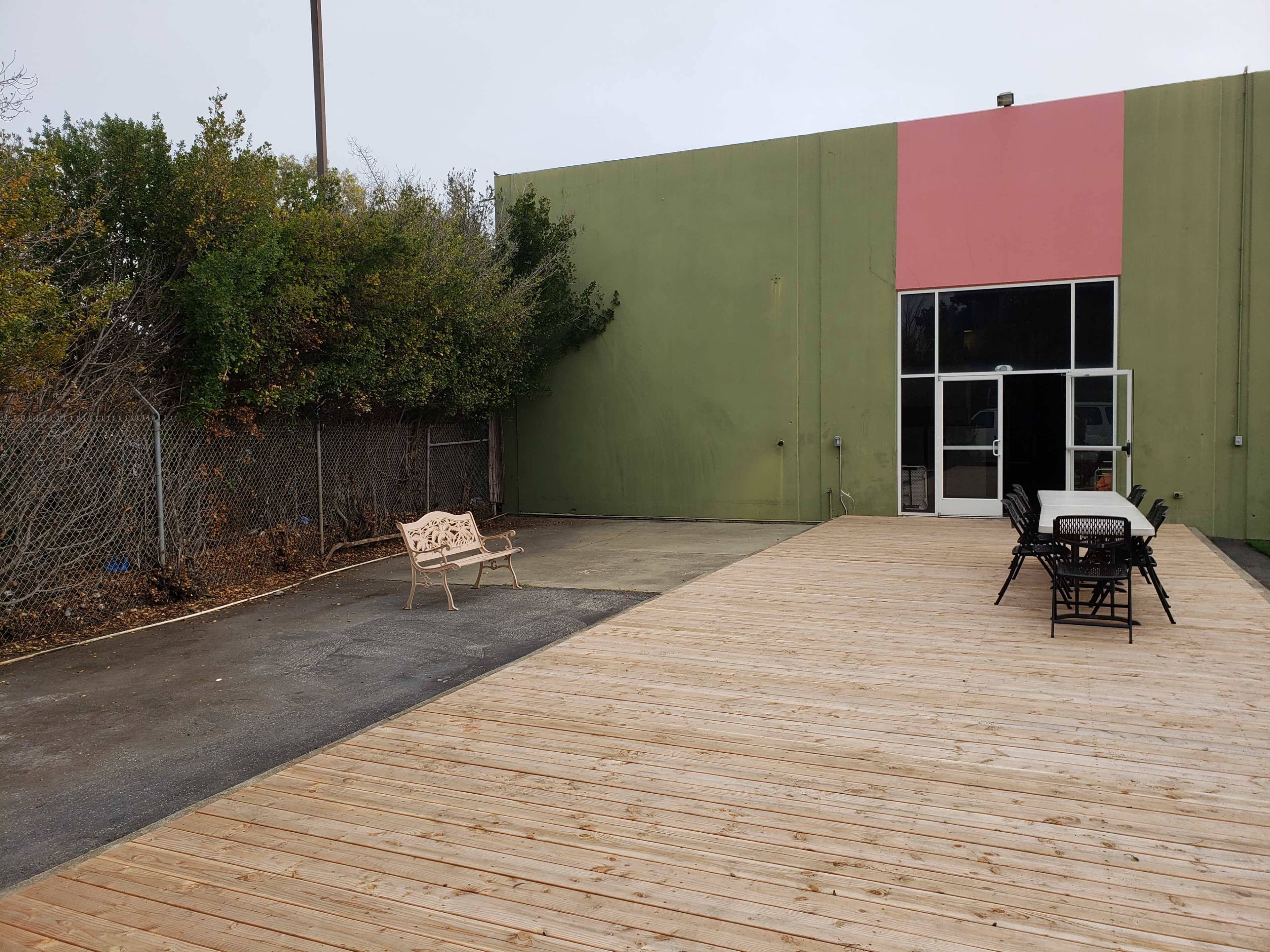 A wooden deck extends from a building with a green wall and a pink section, featuring outdoor seating arrangements and a bench on one side.