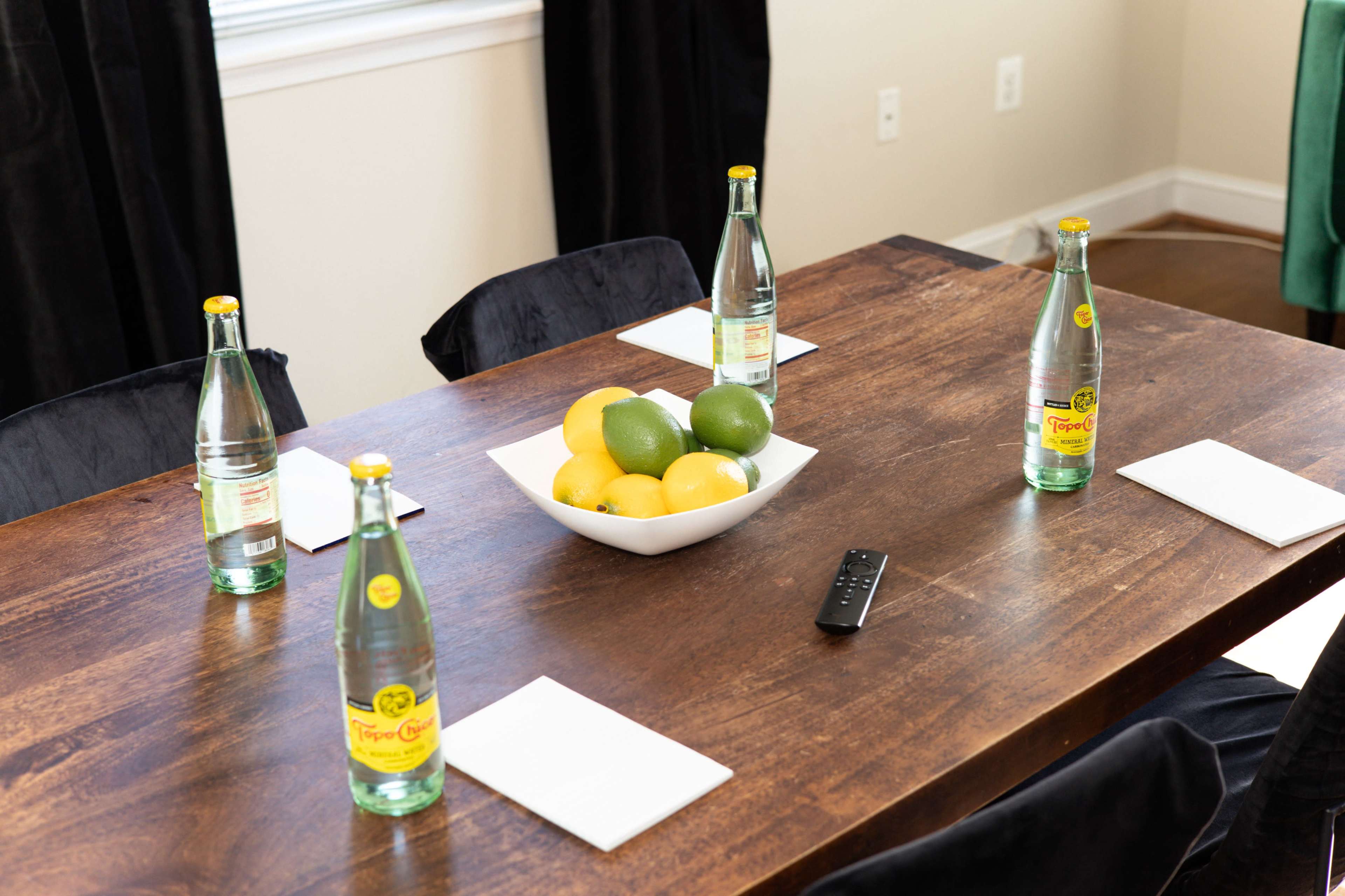 A wooden table is set with sparkling water bottles, coasters, and a bowl of lemons and limes at the center.