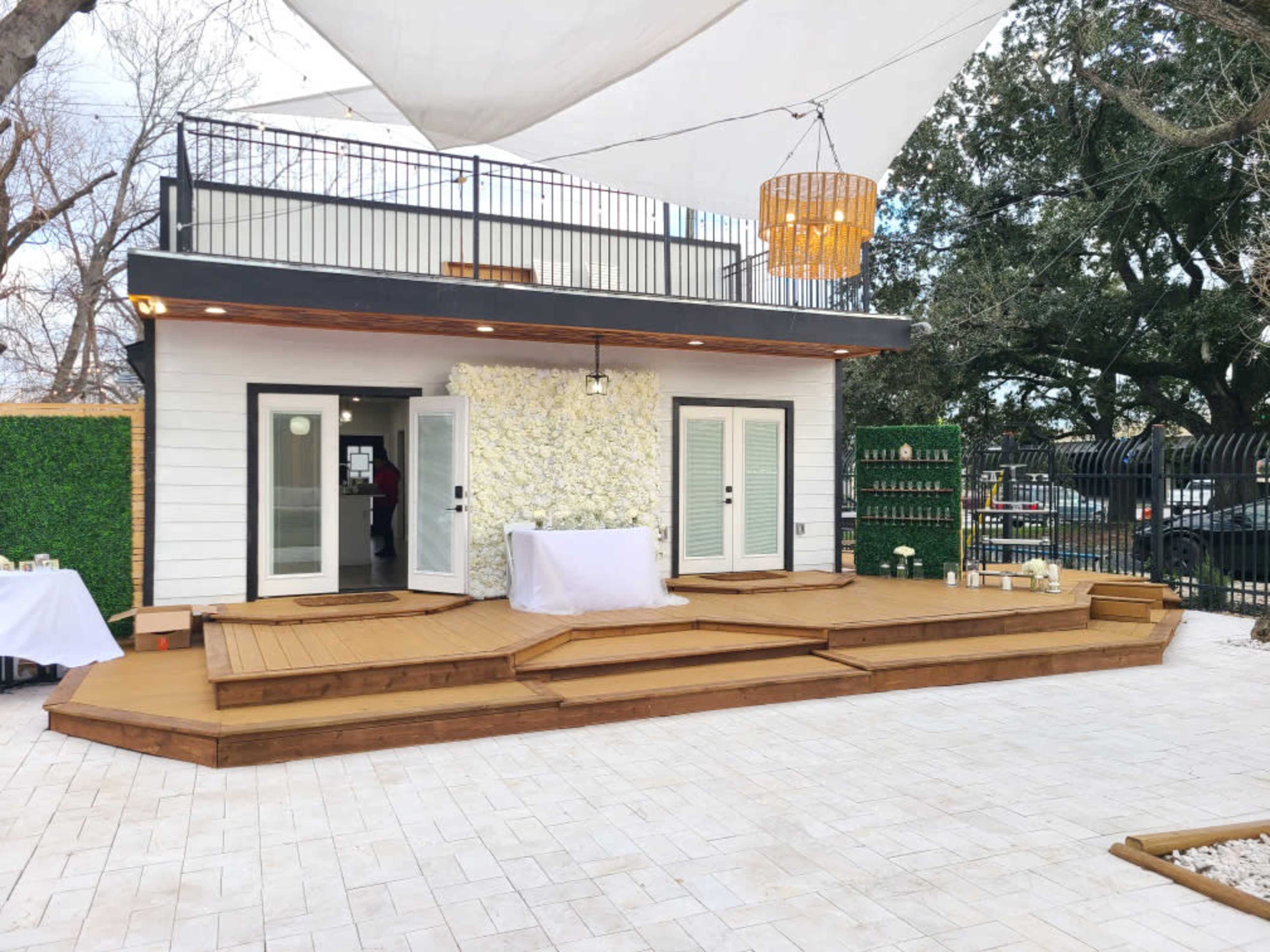 A spacious outdoor deck area with a stage, surrounded by greenery and a modern building featuring large windows and a decorative wall.