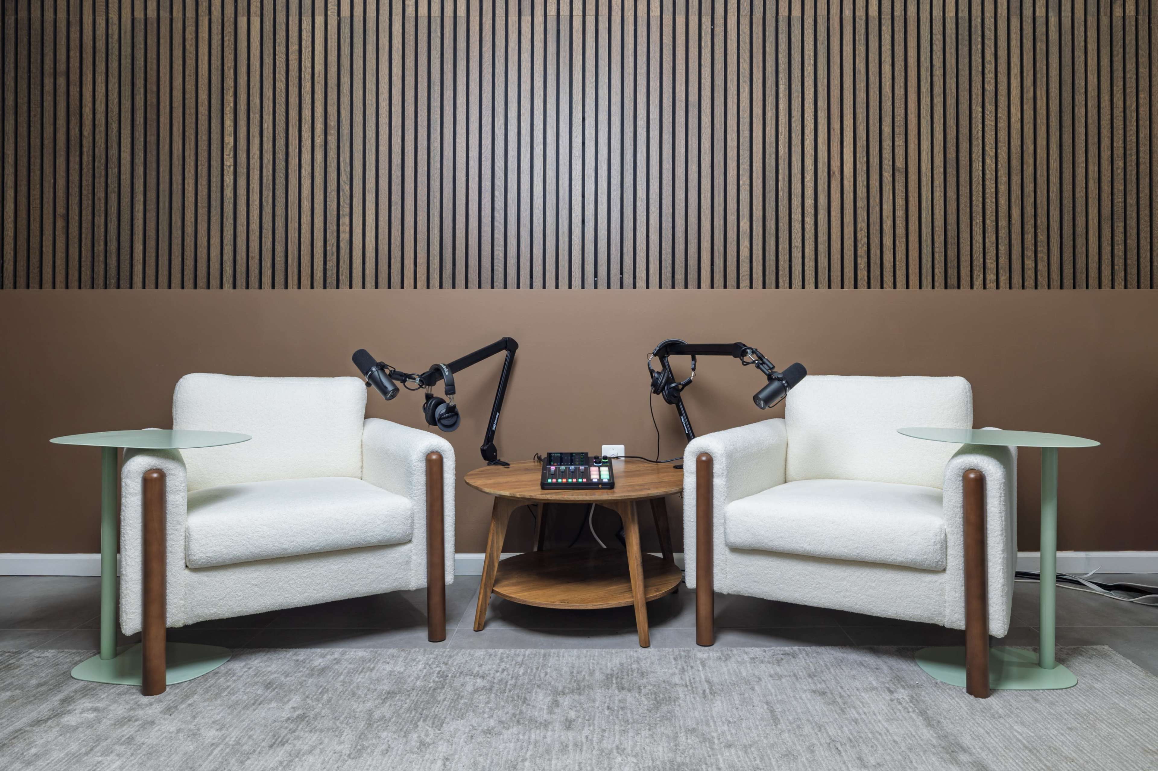 The image shows two white armchairs facing each other with microphone stands attached, separated by a round wooden table and flanked by glass side tables.