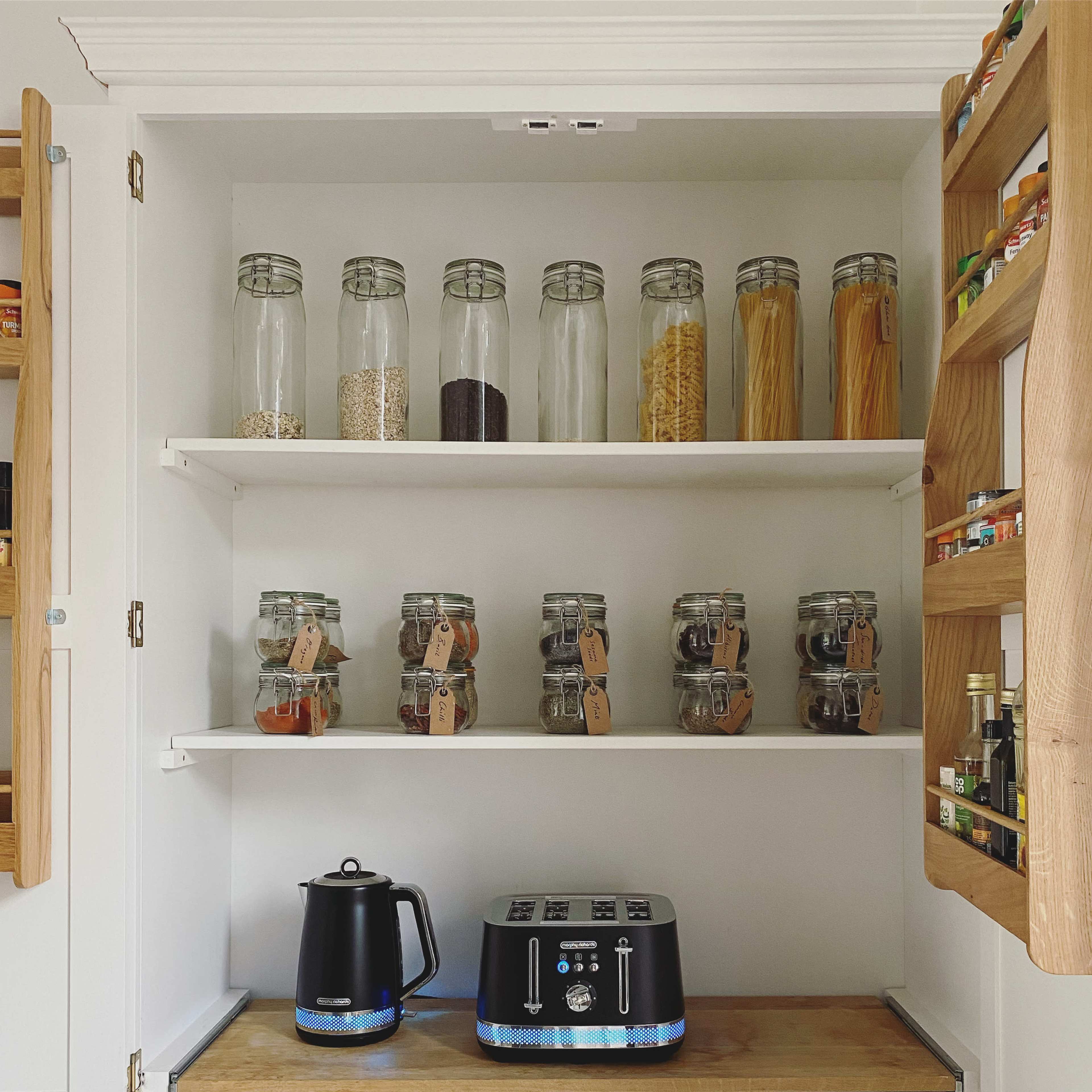 The image shows a neatly organized kitchen pantry with shelves containing glass jars filled with various foods and a kettle on a countertop below.