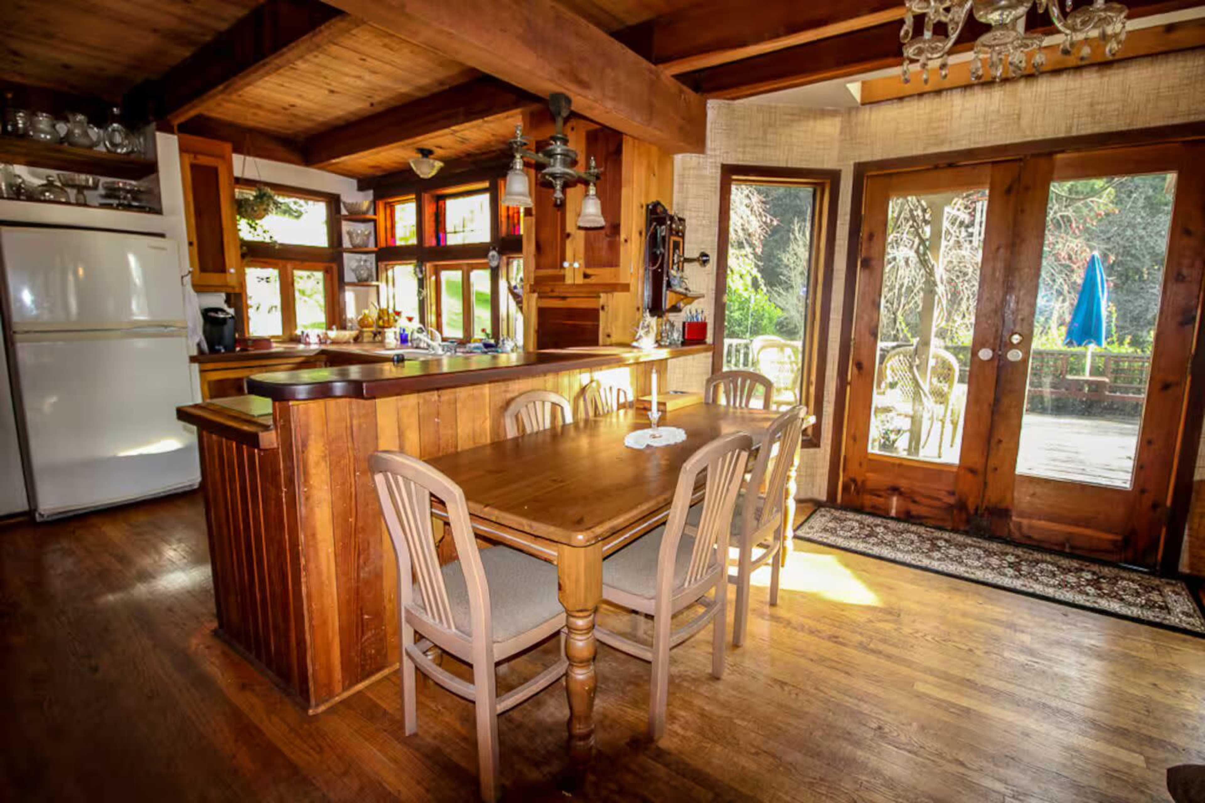 The image shows a rustic kitchen and dining area featuring a wooden table with chairs, a refrigerator, and large glass doors leading to an outdoor space.