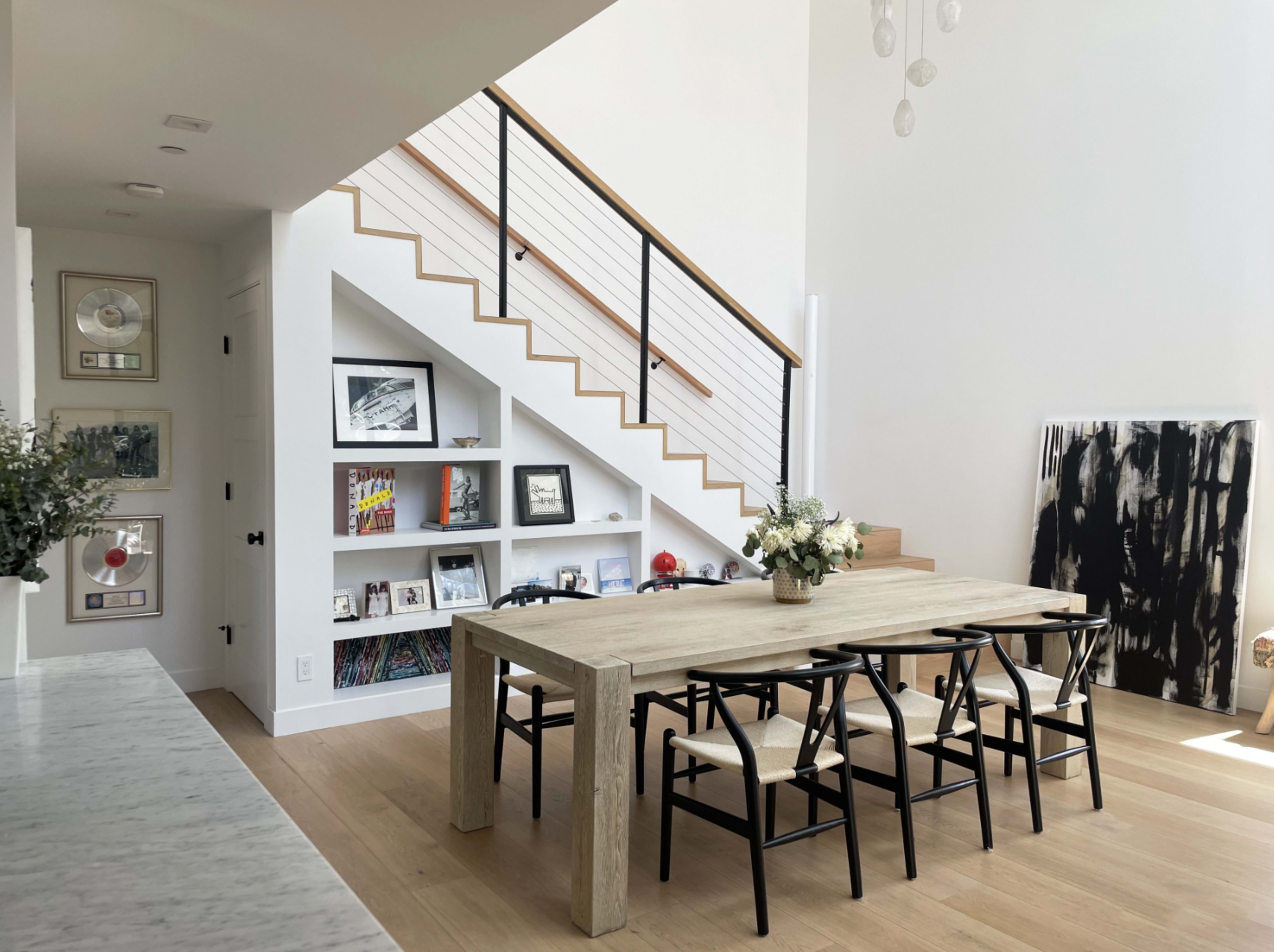 A modern dining area features a long wooden table surrounded by black chairs, with a staircase in the background and shelves displaying framed pictures and decor.