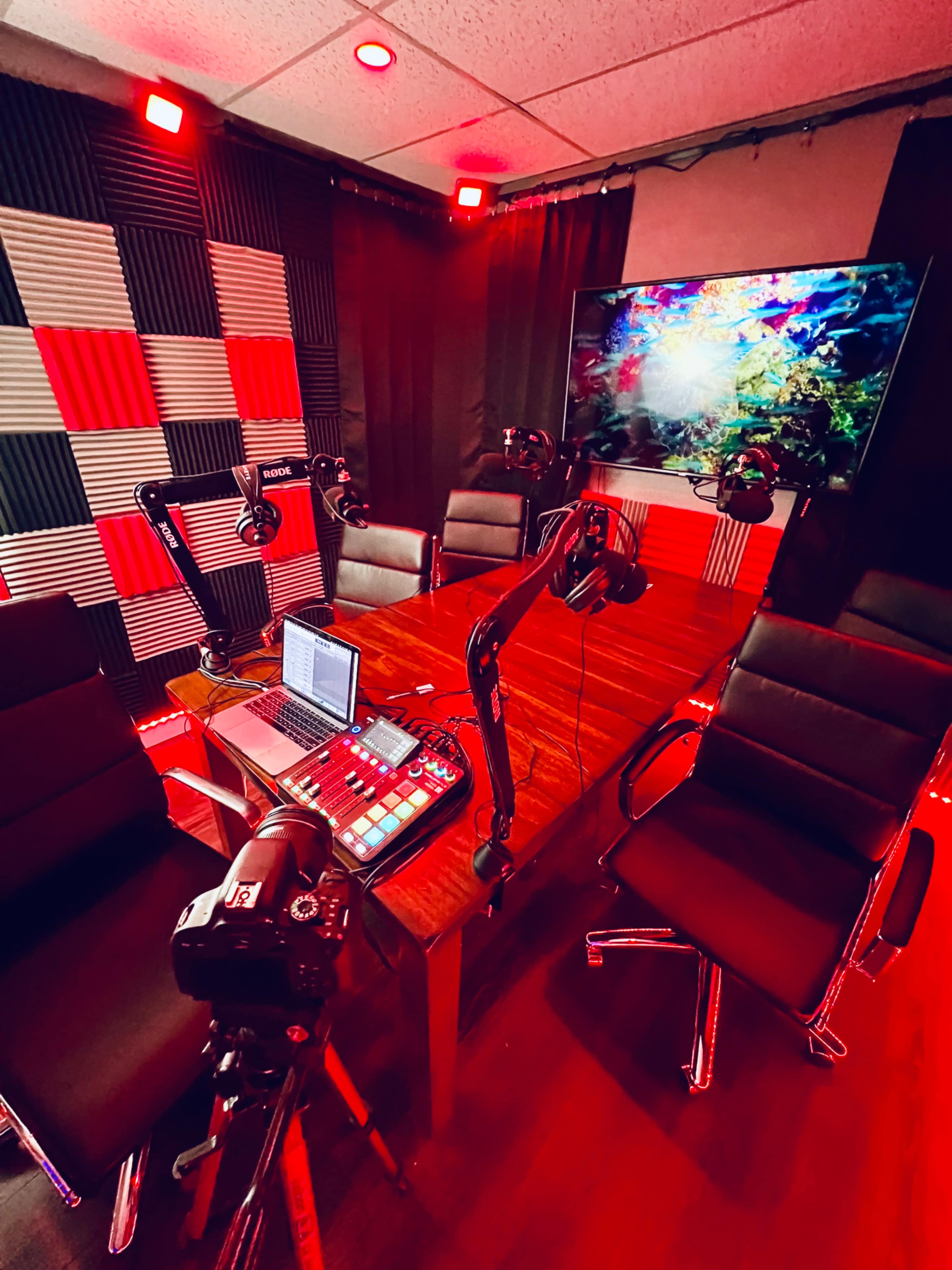 A podcast recording studio with a central wooden table, several black leather chairs, microphone stands, and a television displaying colorful visual content.