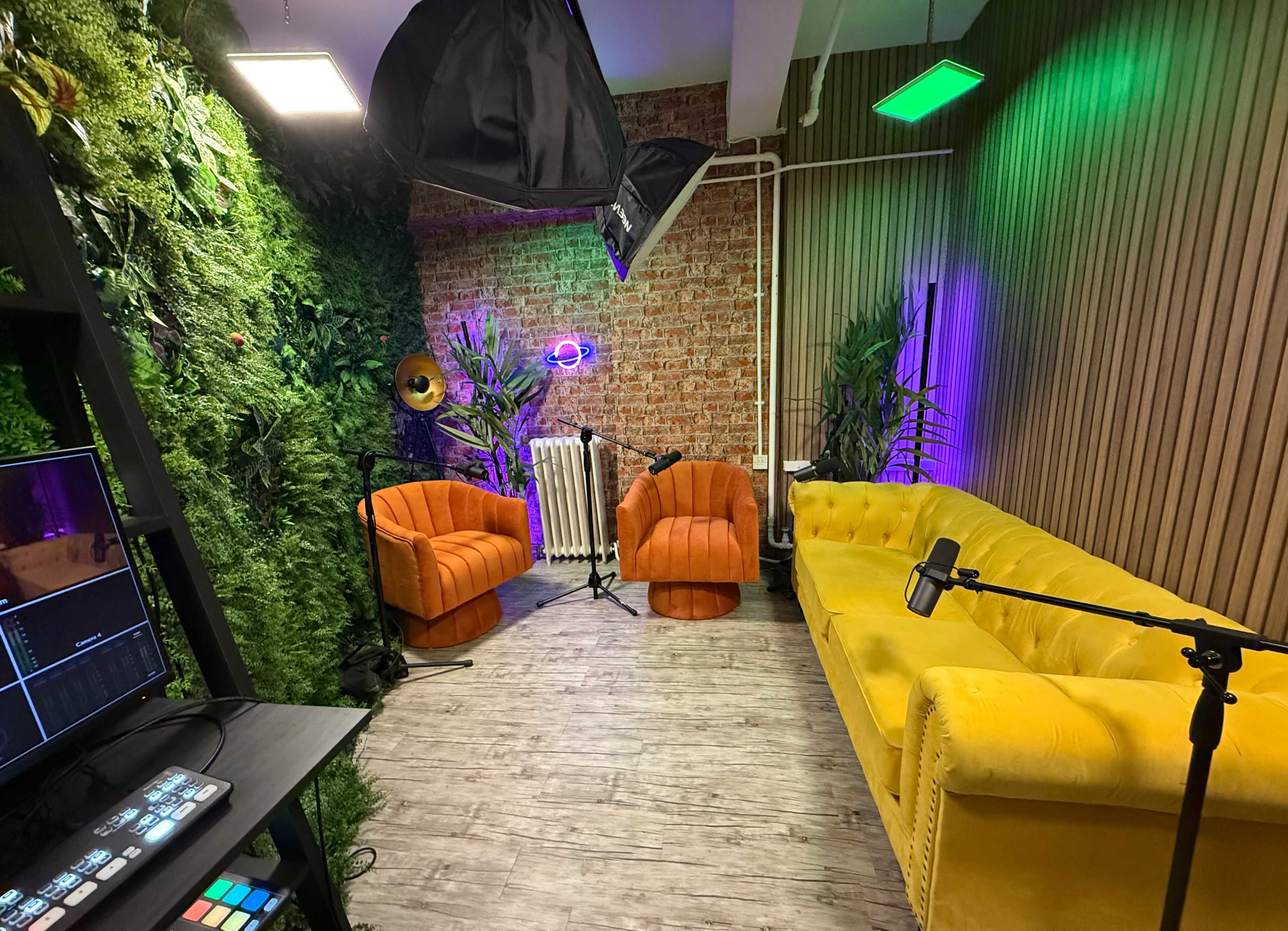 The image shows a brightly colored lounge area with orange and yellow velvet chairs, greenery on the walls, and lighting equipment set up for recording or filming.