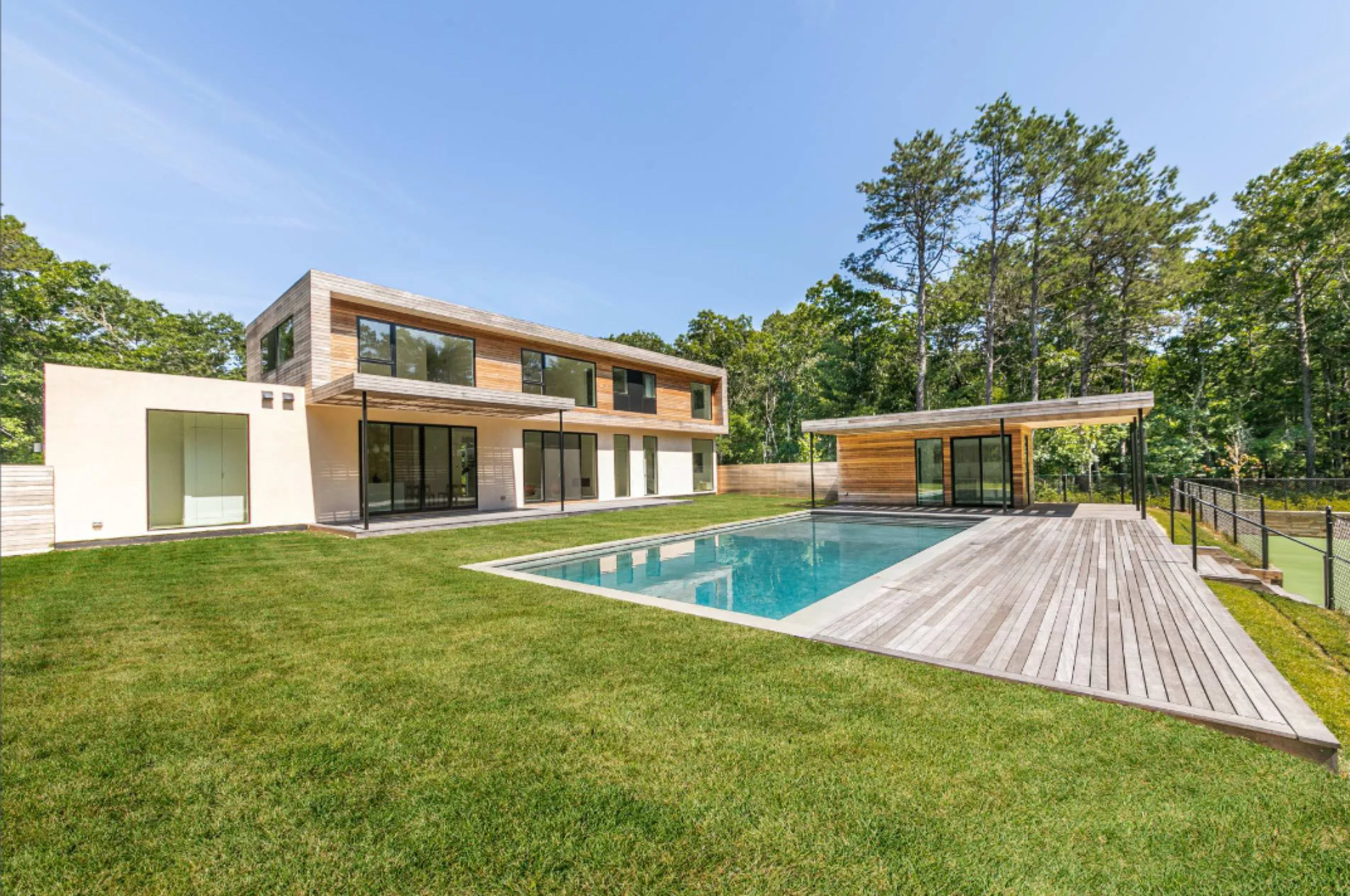 A modern two-story house with large windows and a wooden deck surrounds a rectangular swimming pool in a grassy yard bordered by trees.