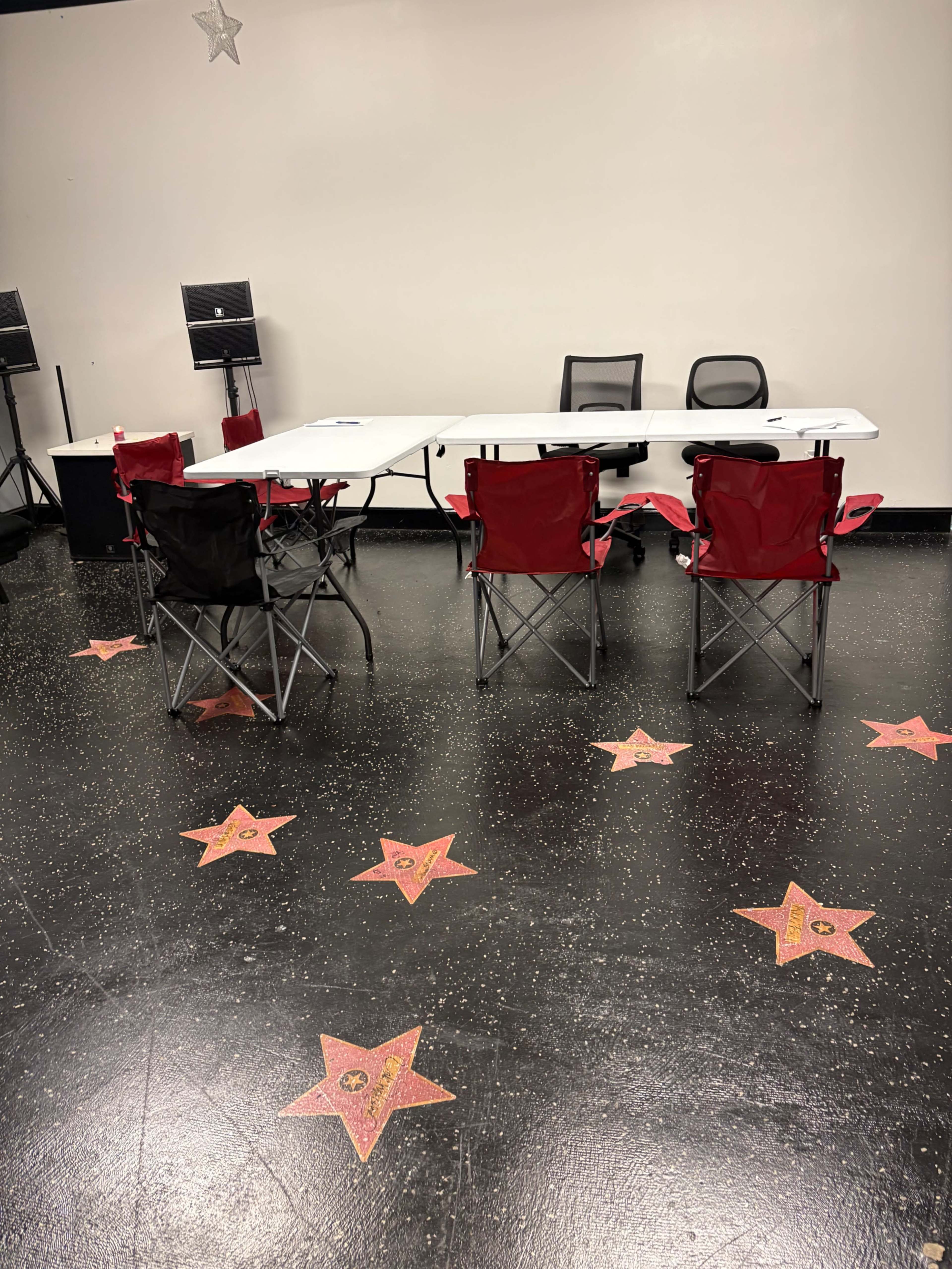 The image shows a blank room featuring a long table with black and red folding chairs, surrounded by star-shaped decorations on the floor.