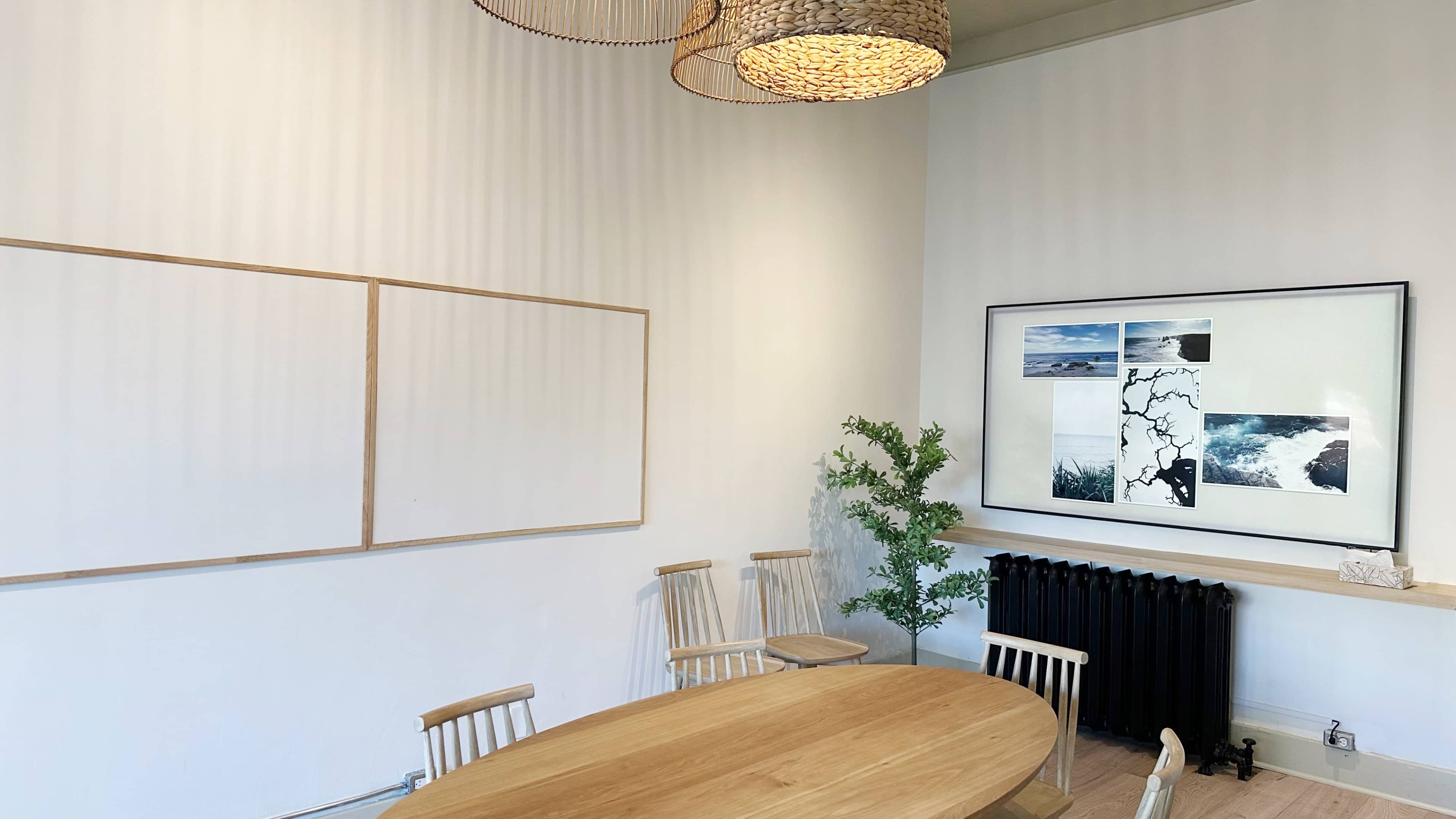A minimalist meeting room features a round wooden table, several wooden chairs, a large framed collage on the wall, and a radiator beneath a plant.