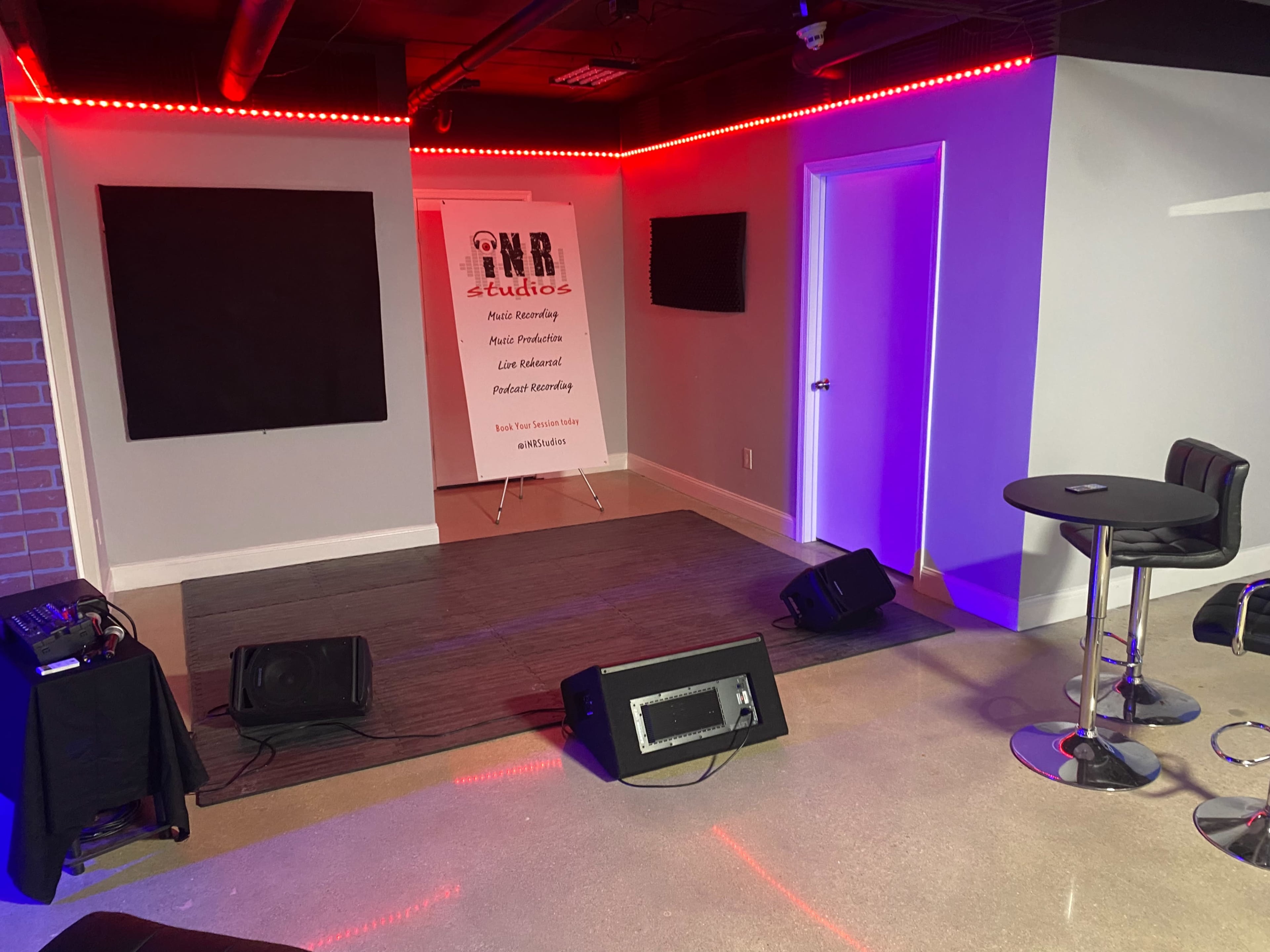 The image shows a small music studio with a stage area, featuring a microphone stand, sound equipment, and a backdrop with text, surrounded by colorful lighting and seating.