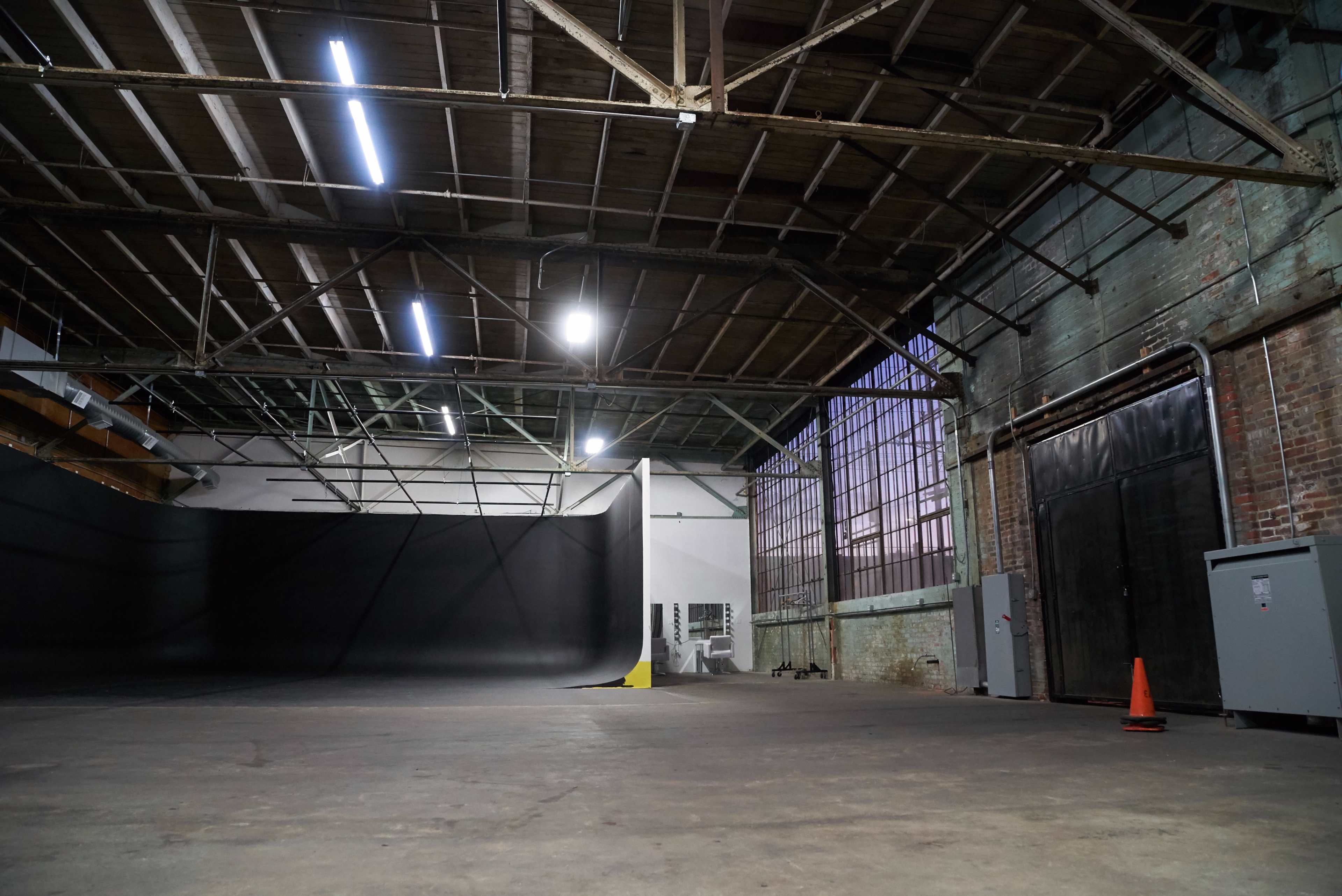 The image shows an empty warehouse space with high ceilings, exposed beams, and a large, black backdrop facing a window.