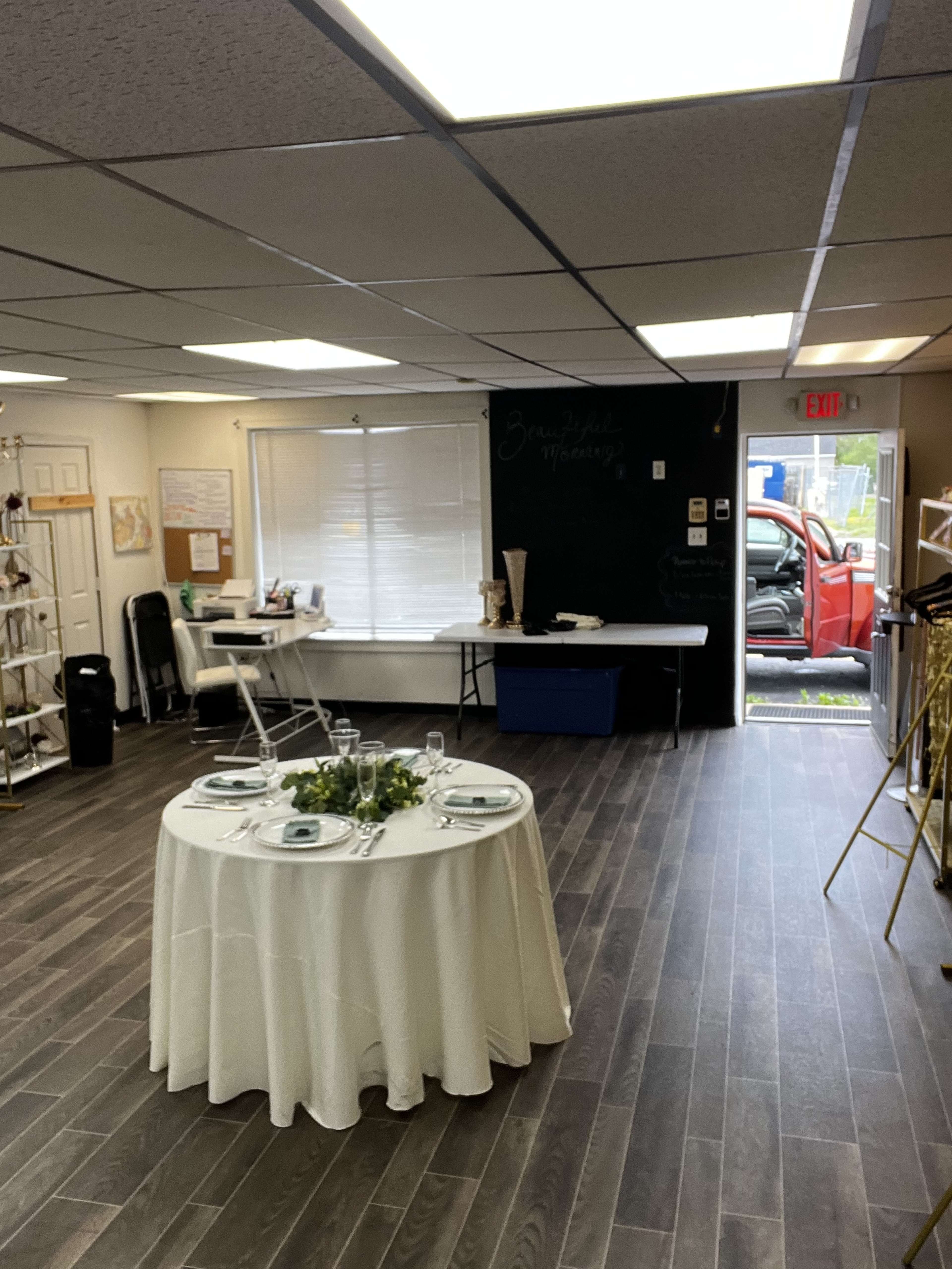 The image shows a small event space featuring a round dining table set with plates and cutlery, a black chalkboard, and an open door leading to a vehicle outside.