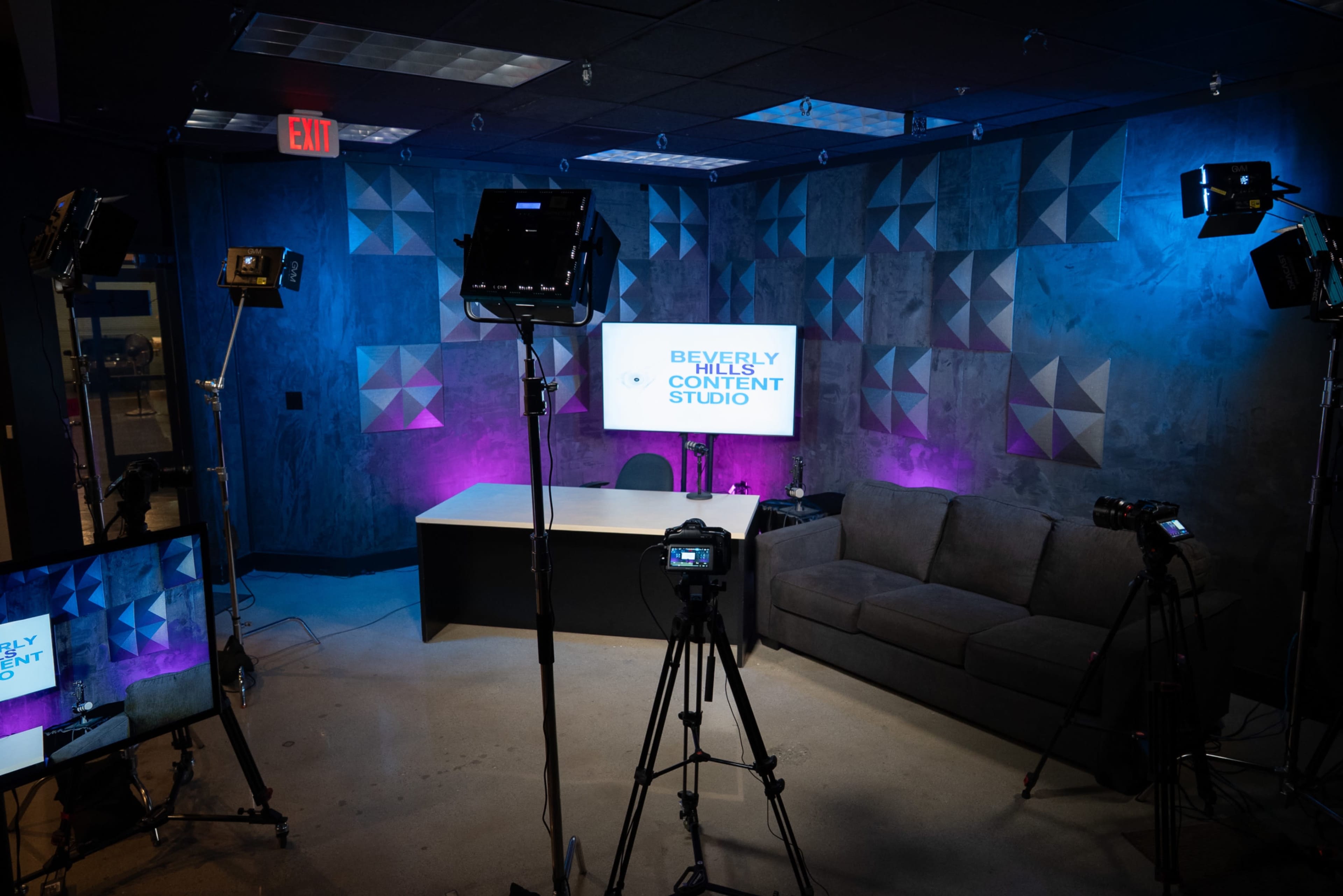 The image shows a content studio with a modern design featuring a gray wall, a desk, a couch, and several video cameras set up for recording.