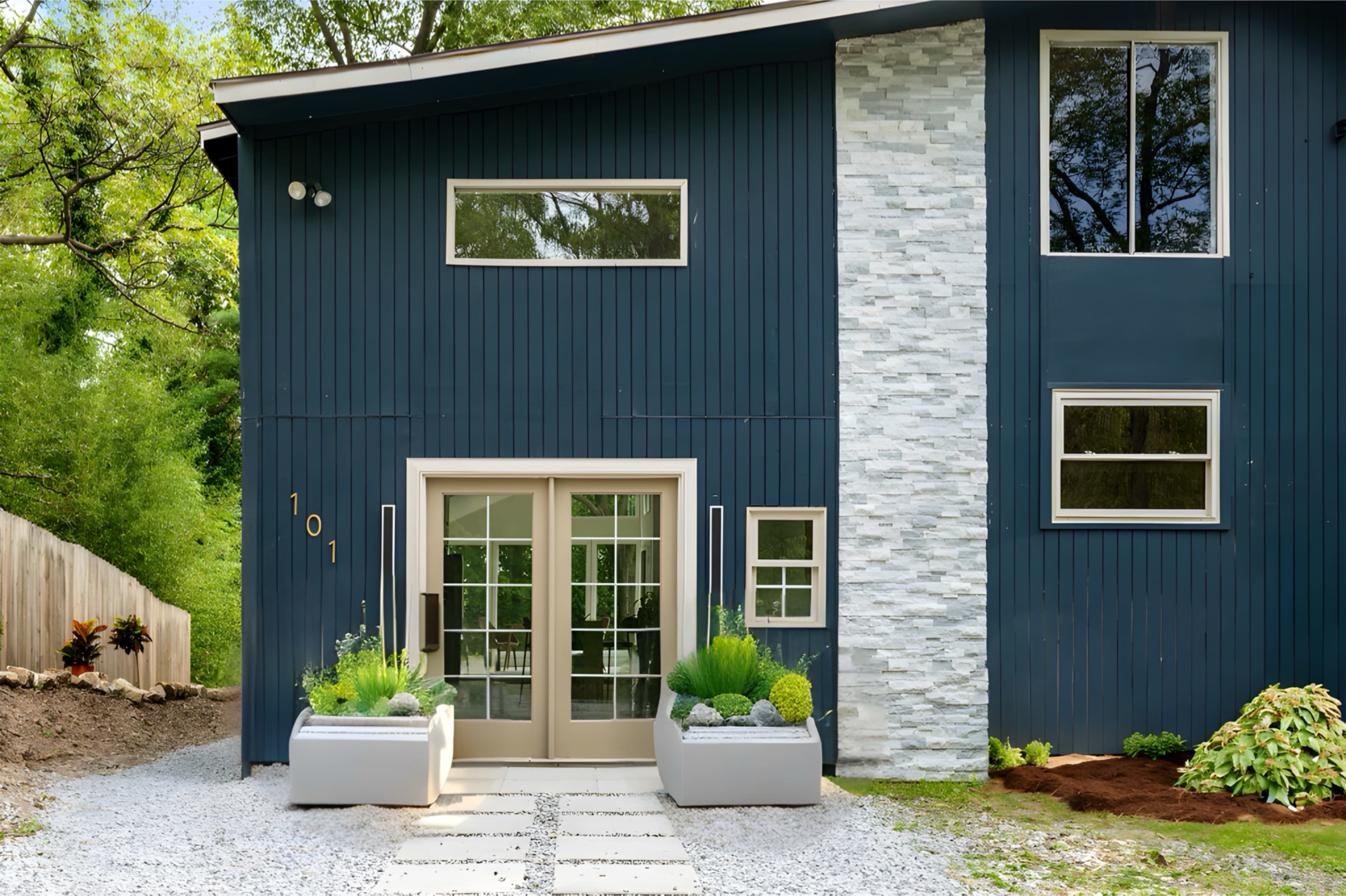 The image shows a modern blue home with large windows, a stone accent wall, and a minimalist entrance framed by two planters.
