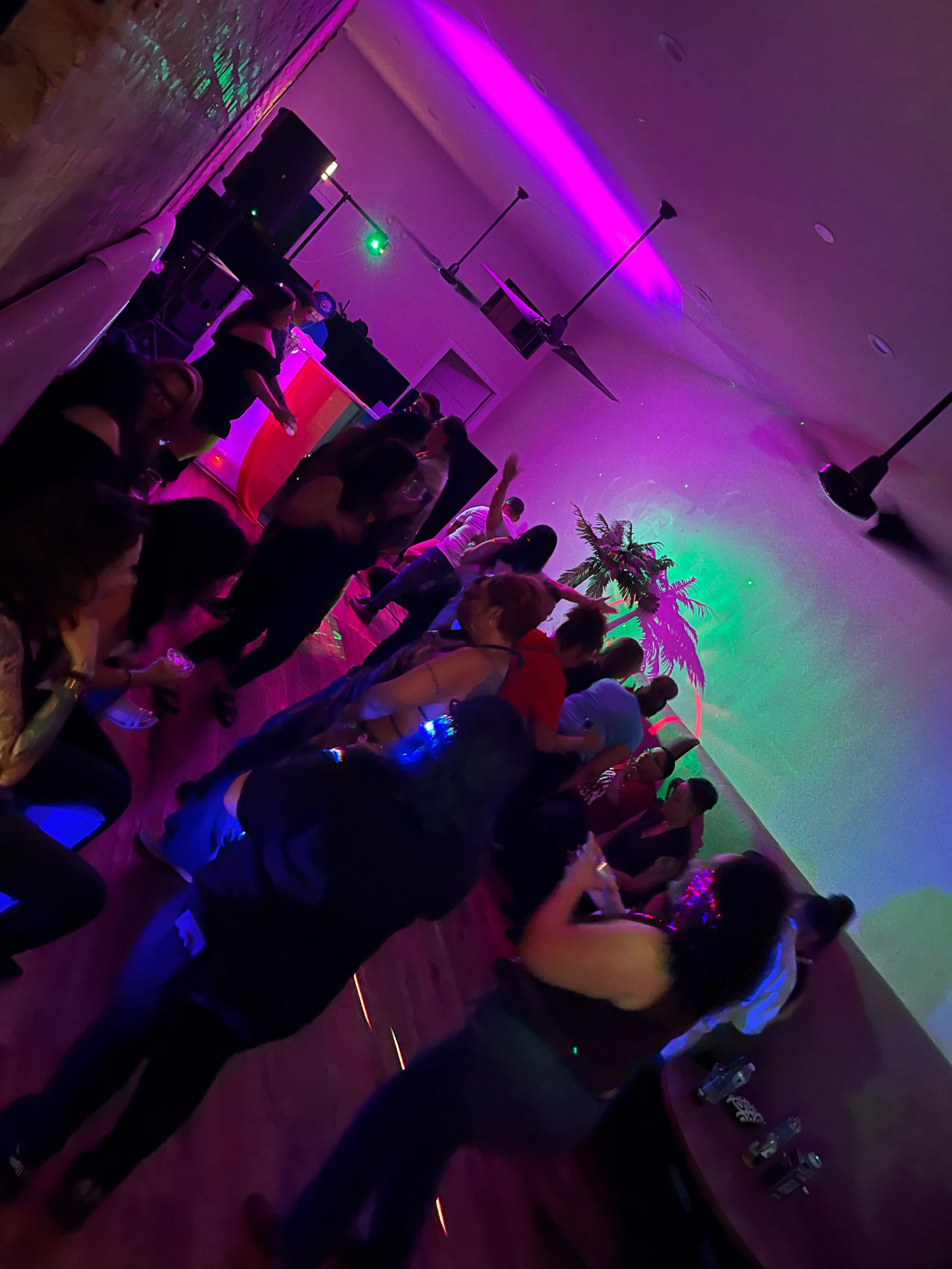 A group of people dance in a dimly lit room with colorful lights and a DJ setup in the background.