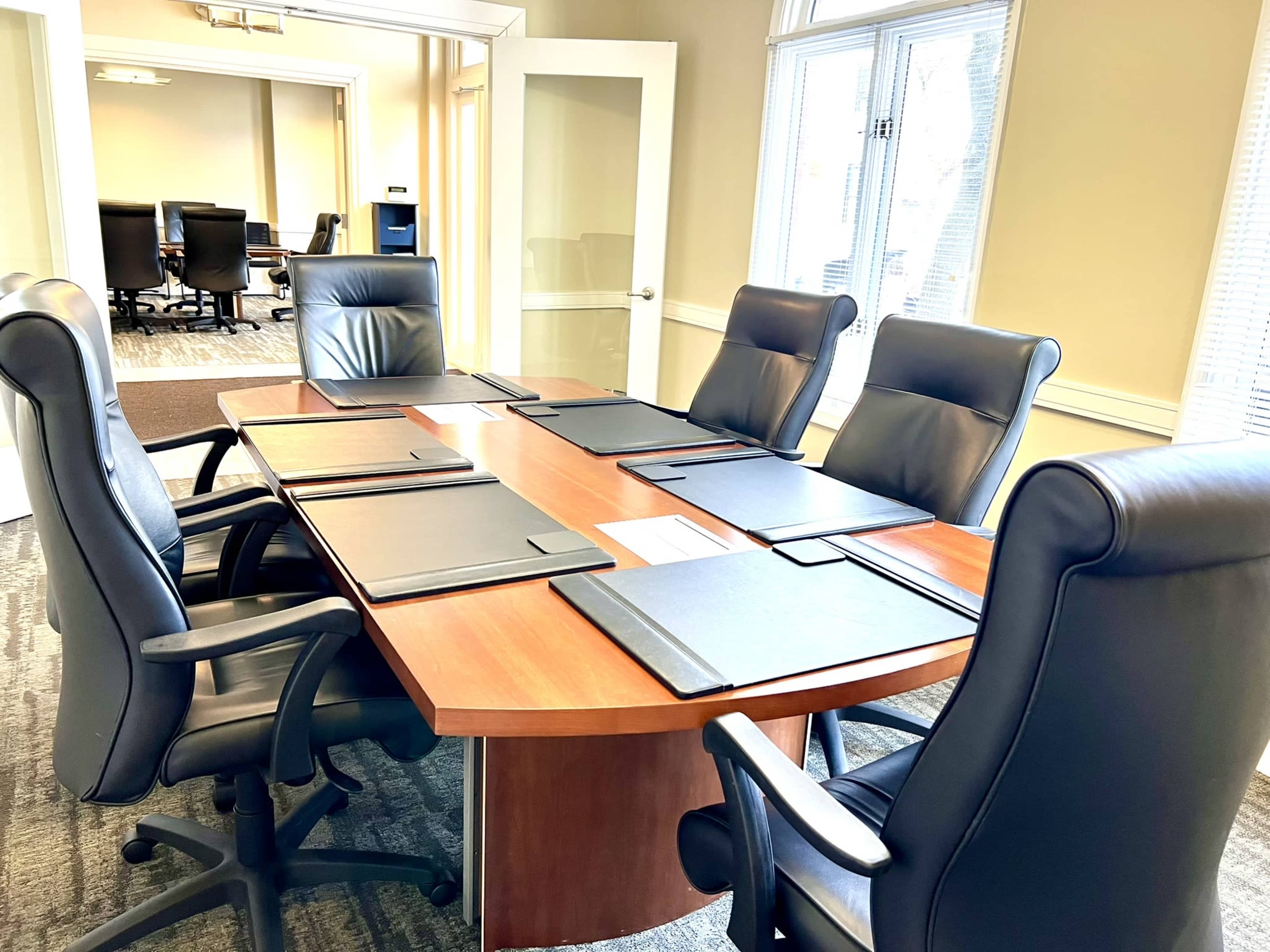 A conference room features a large wooden table surrounded by black leather chairs, with several empty folders placed on the table.