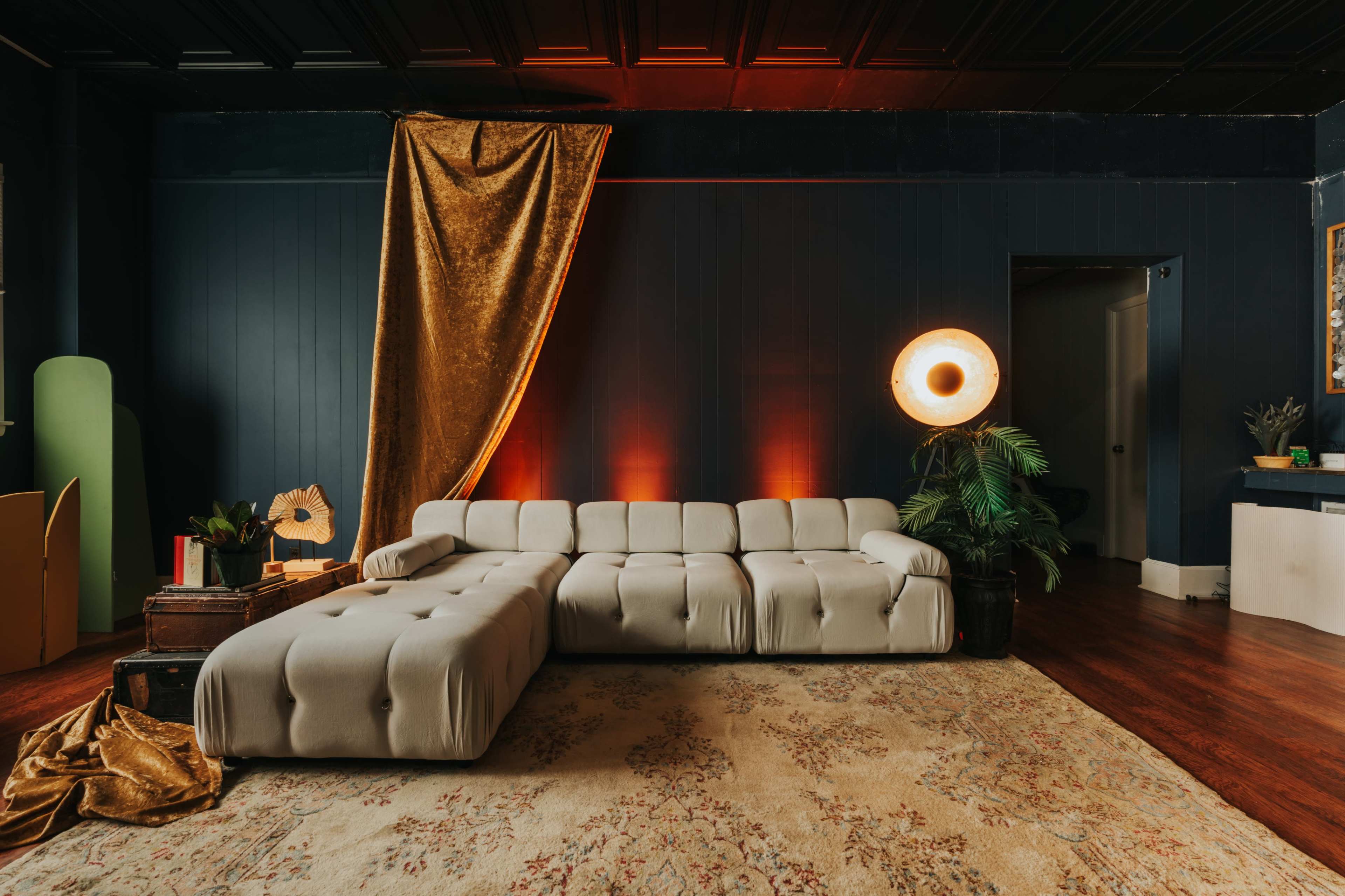 A modern living room featuring a plush sectional sofa, a large area rug, and warm lighting against a dark wall, complemented by decorative plants and a golden drapery.