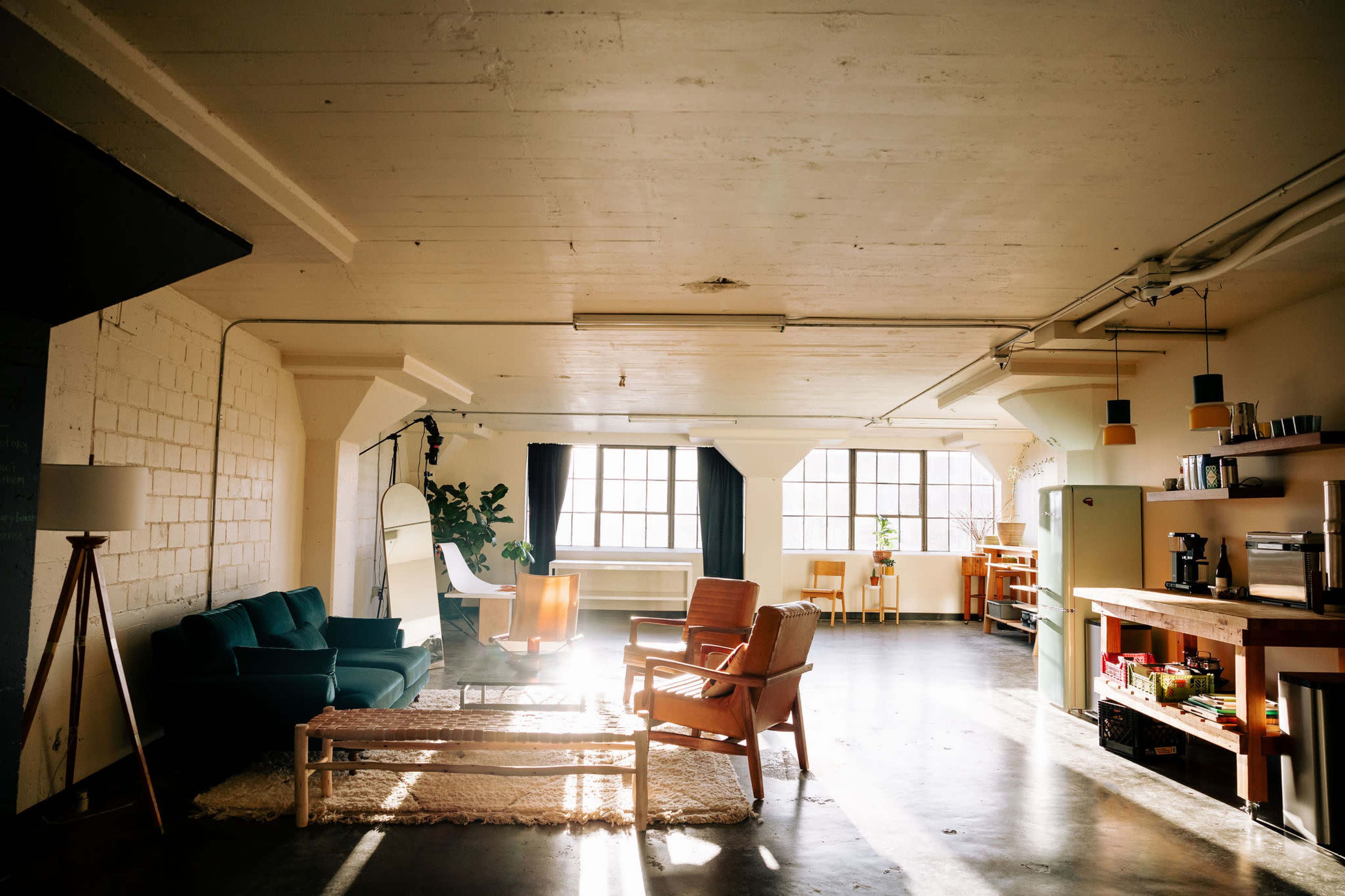 A spacious, bright loft-style room with large windows, featuring a seating area with green and brown chairs, a wooden coffee table, and an open kitchen space.