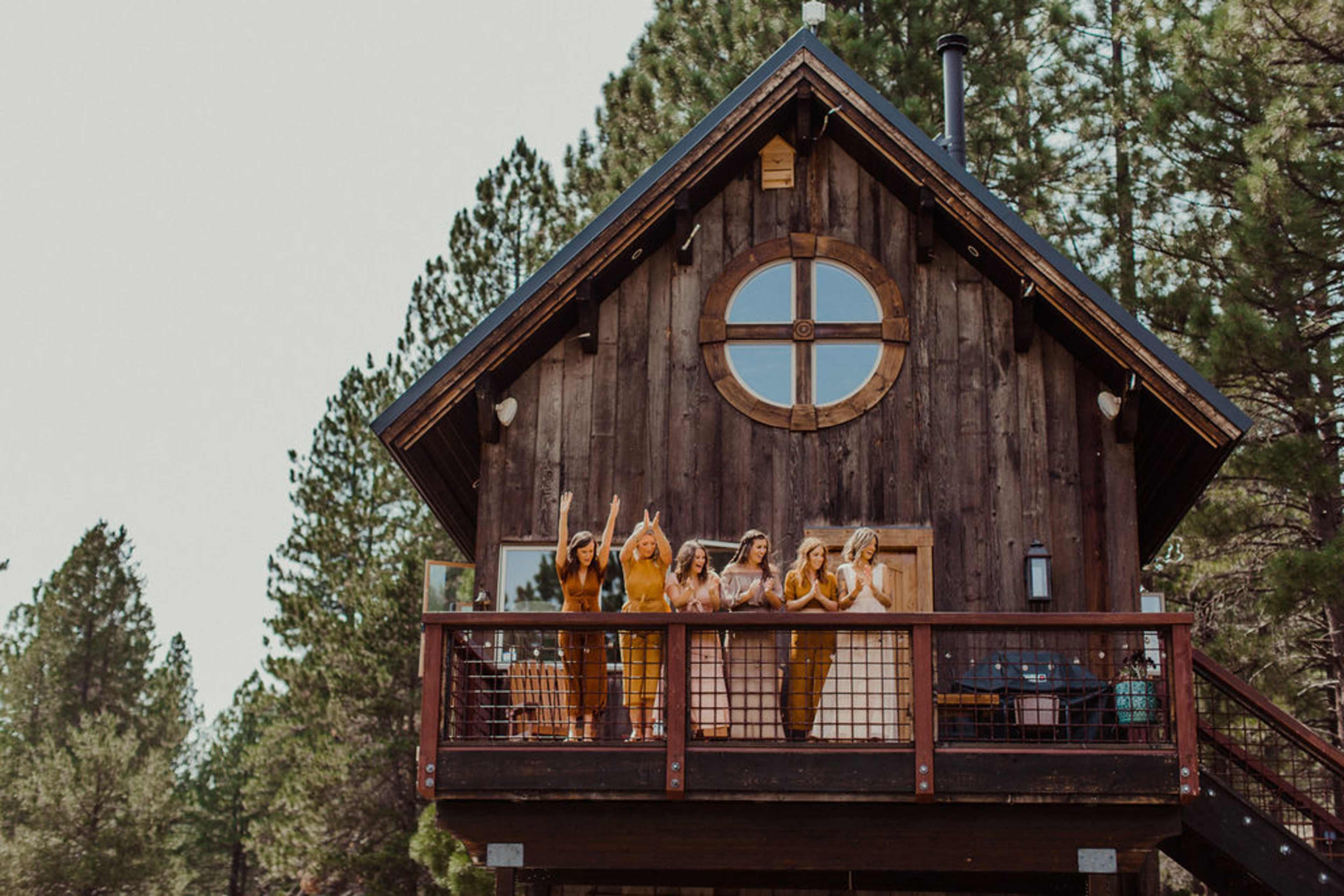 A group of women in matching outfits cheer from the balcony of a wooden cabin surrounded by trees.