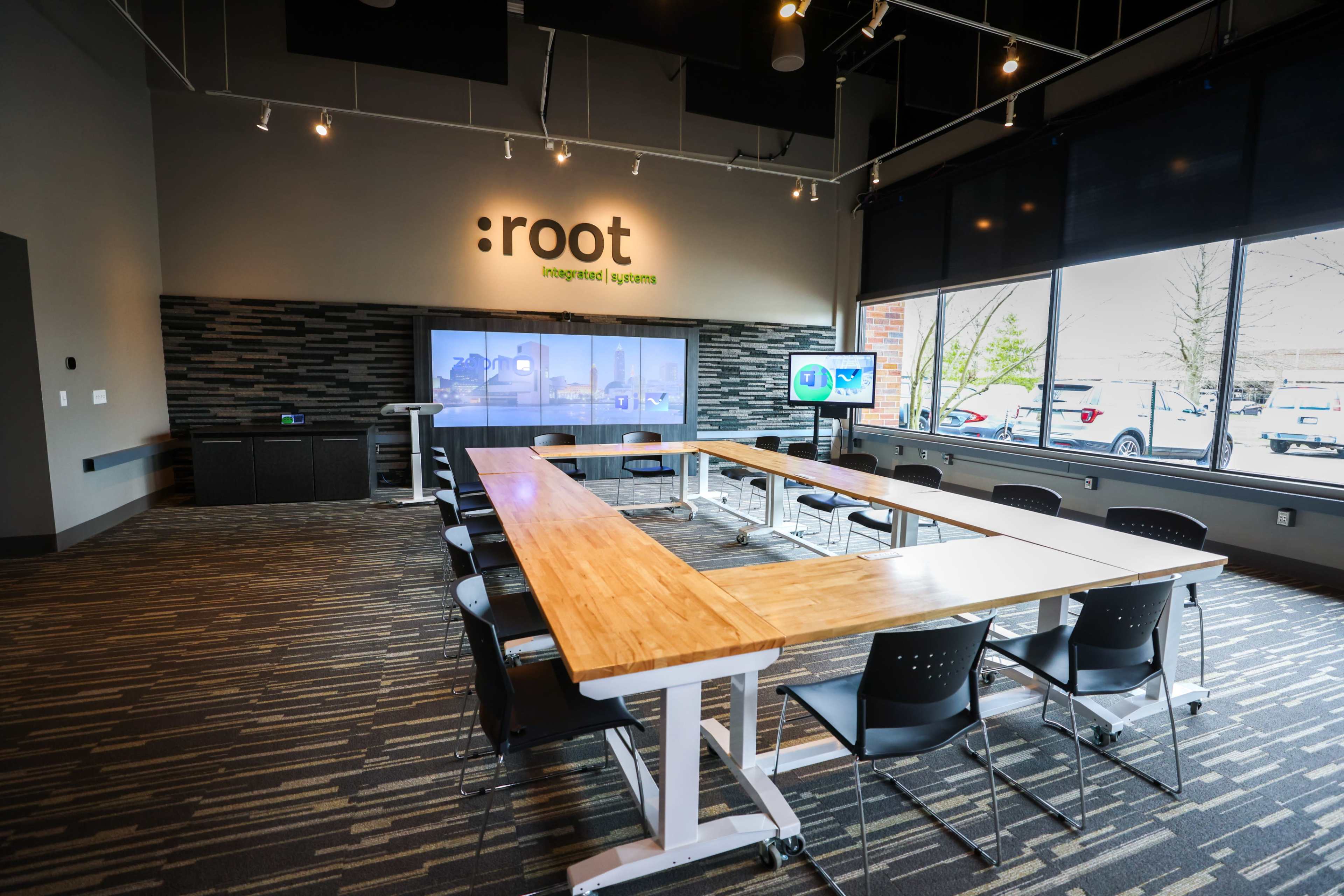 A modern conference room with a large wooden table, black chairs, and a screen displaying a presentation on one wall.