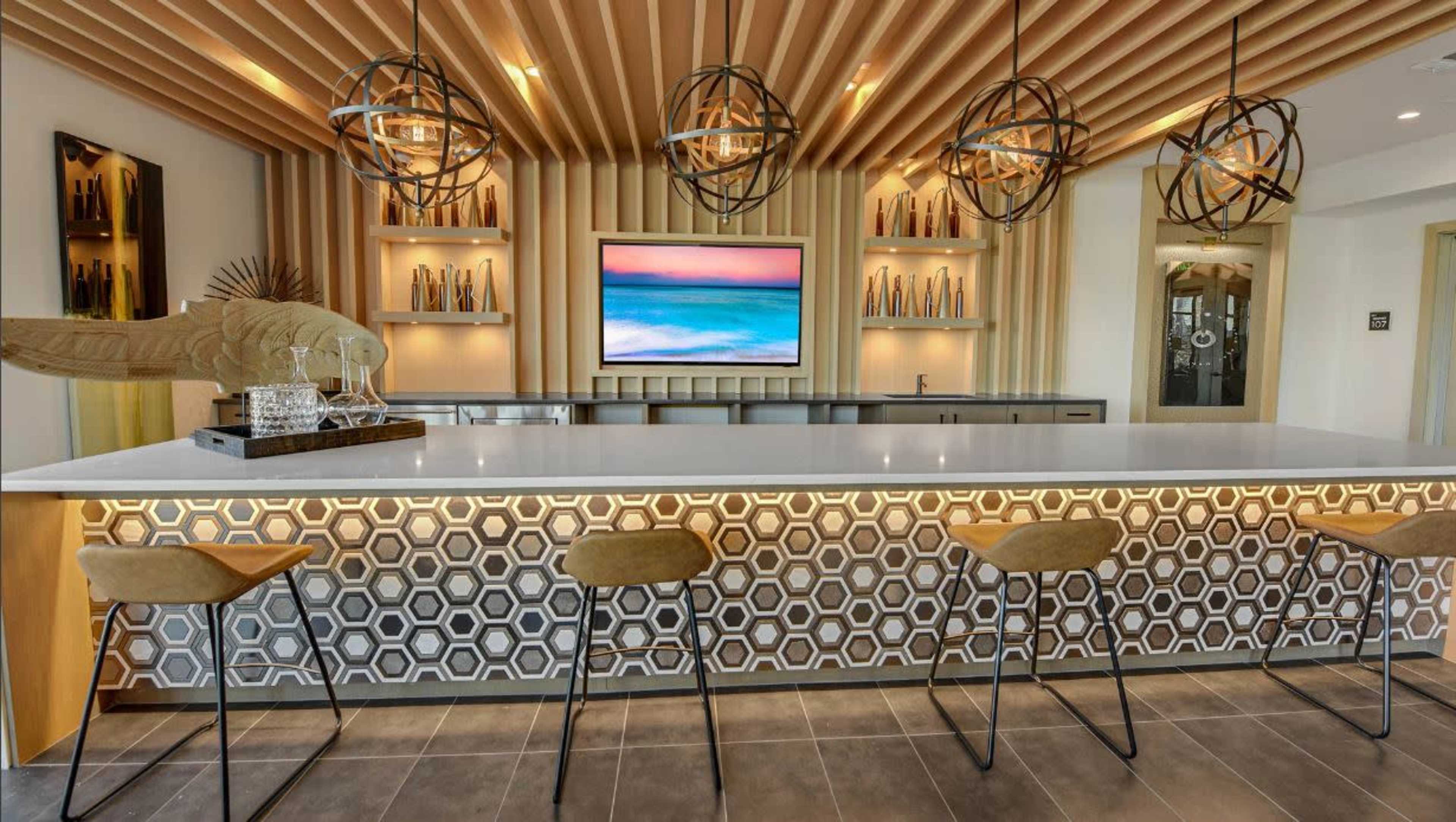 A modern bar area with a white countertop, geometric patterned front, and pendant lighting above, complemented by a large screen displaying a scenic view.
