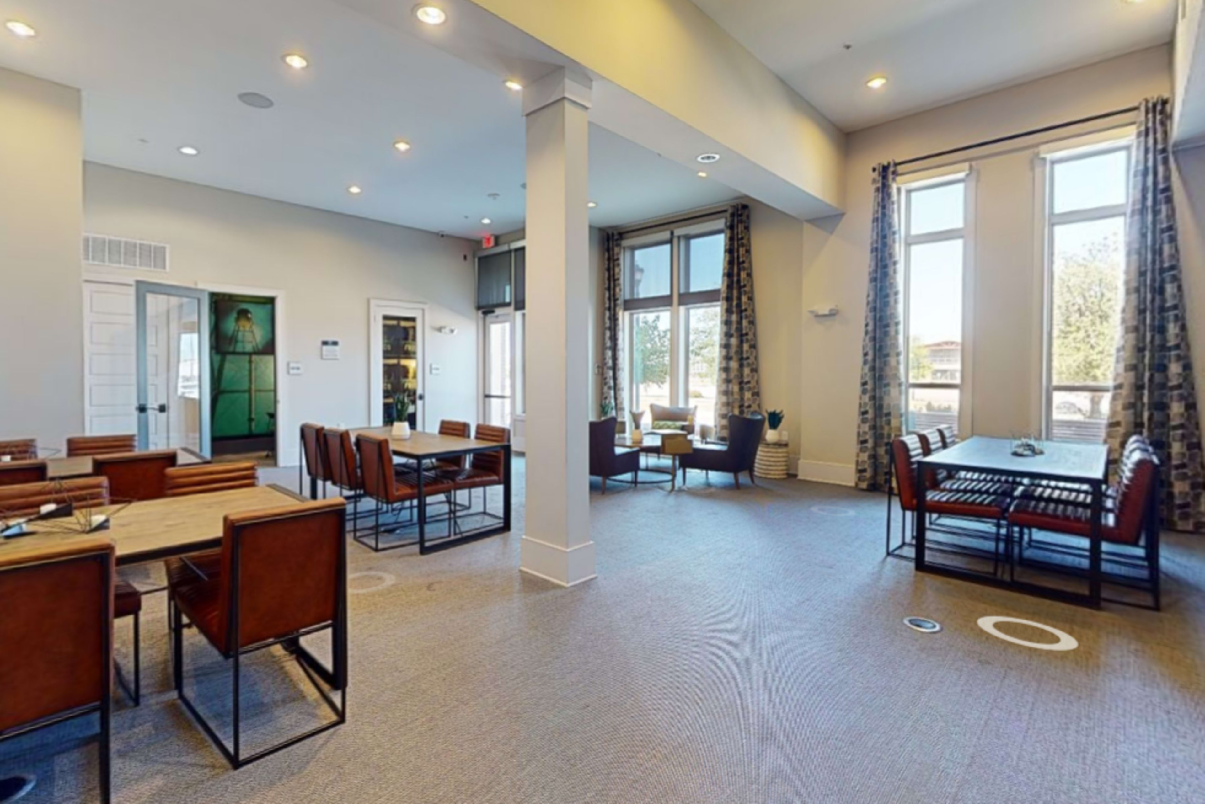 The image shows a spacious indoor dining area with tables and chairs arranged around the room, featuring large windows that allow natural light to enter.