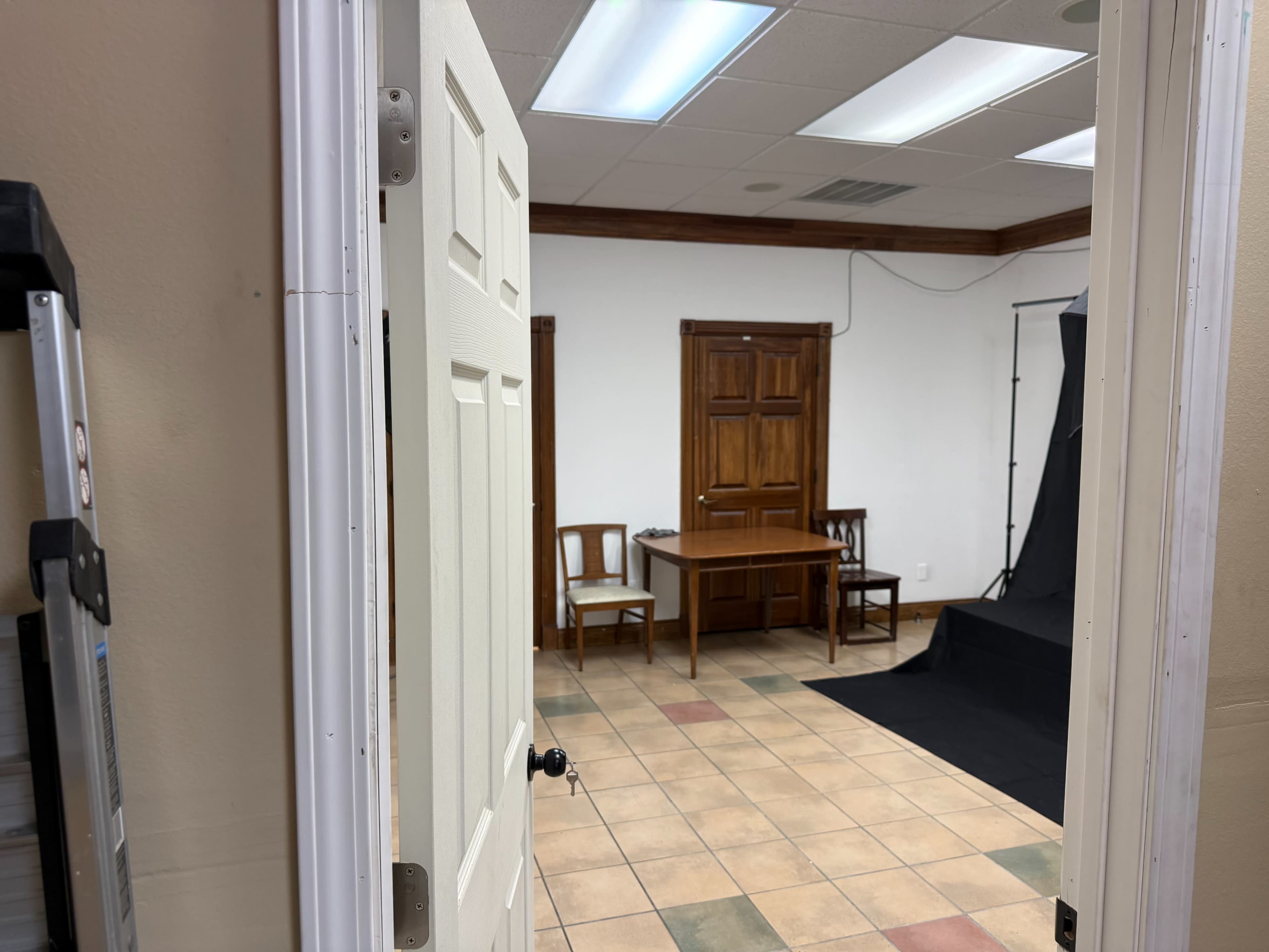 The image shows an interior room with two wooden chairs and a table, a black backdrop, and an open door leading into the space.