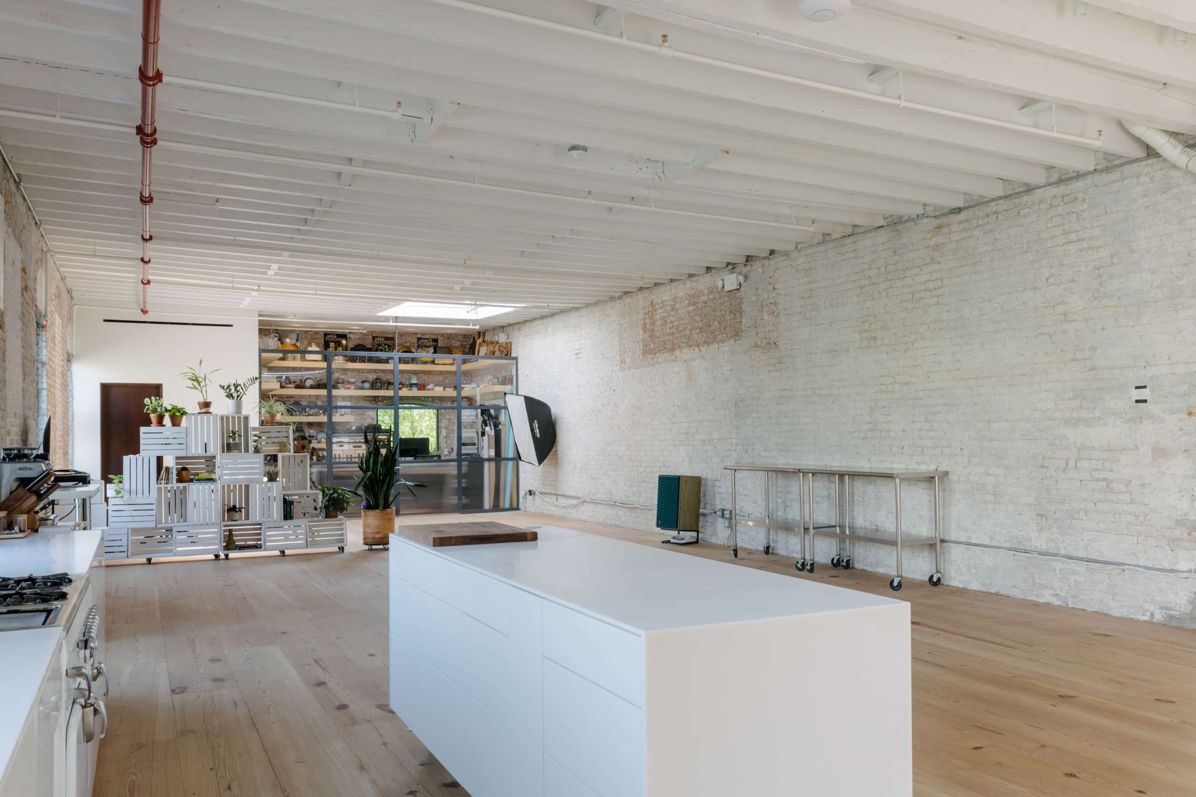 The image shows a spacious kitchen with a central white island, wooden flooring, and exposed brick walls, featuring shelving filled with plants and kitchen items in the background.