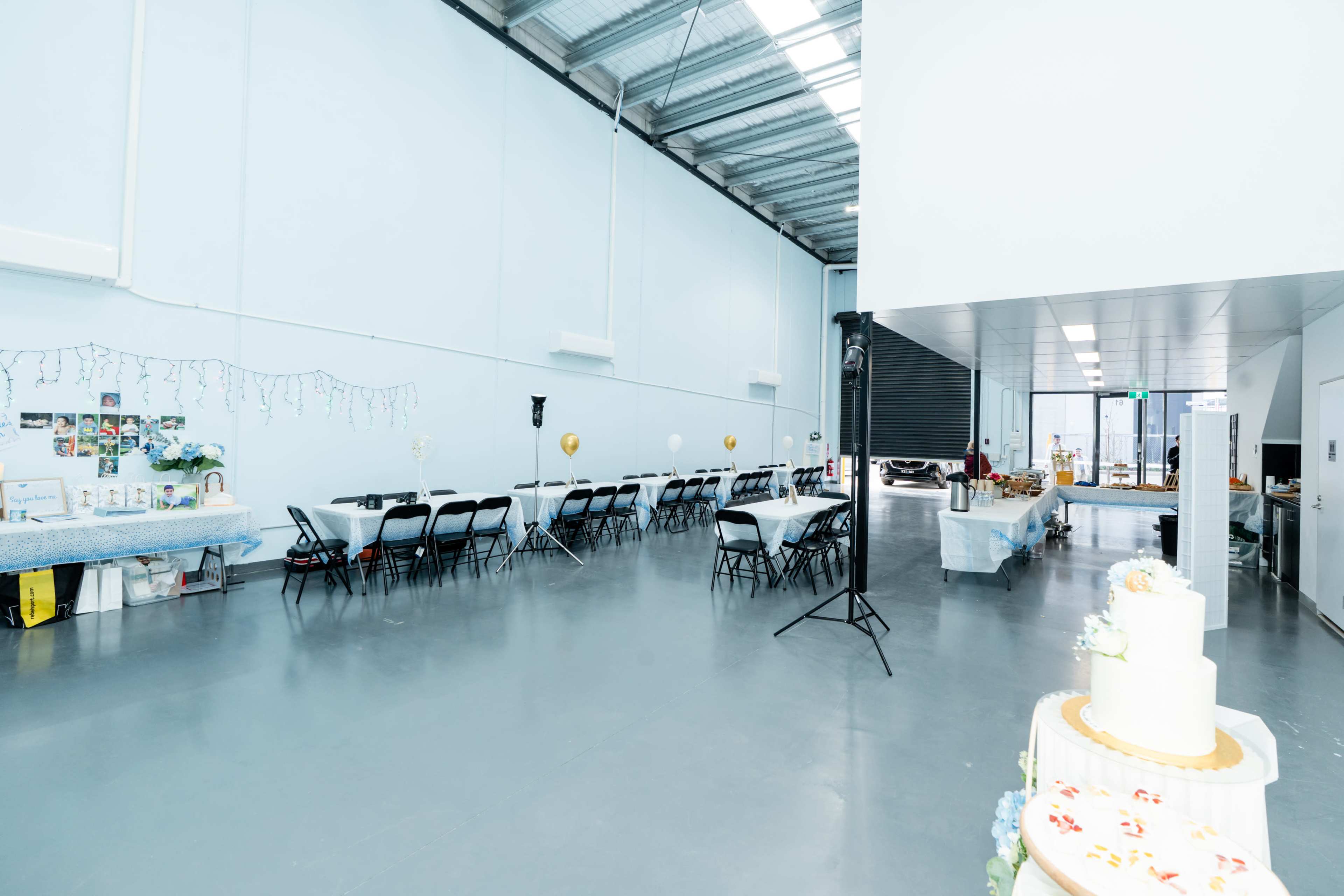 The image shows an empty indoor event space with tables and chairs arranged along one side, and decorations lining the wall.