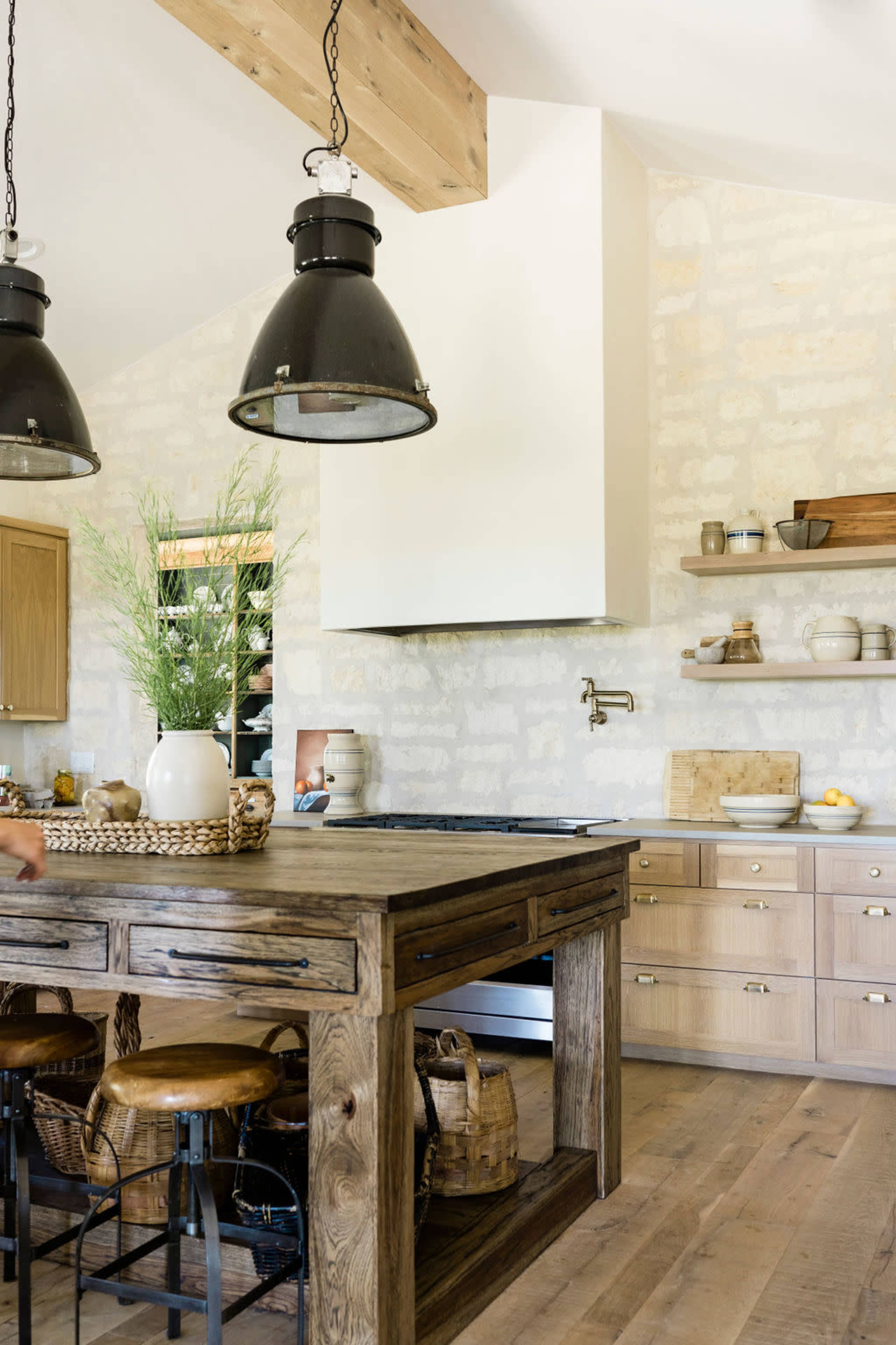 The kitchen features a large wooden island with round stools, industrial pendant lights, and a simple decor against a stone and wood backdrop.