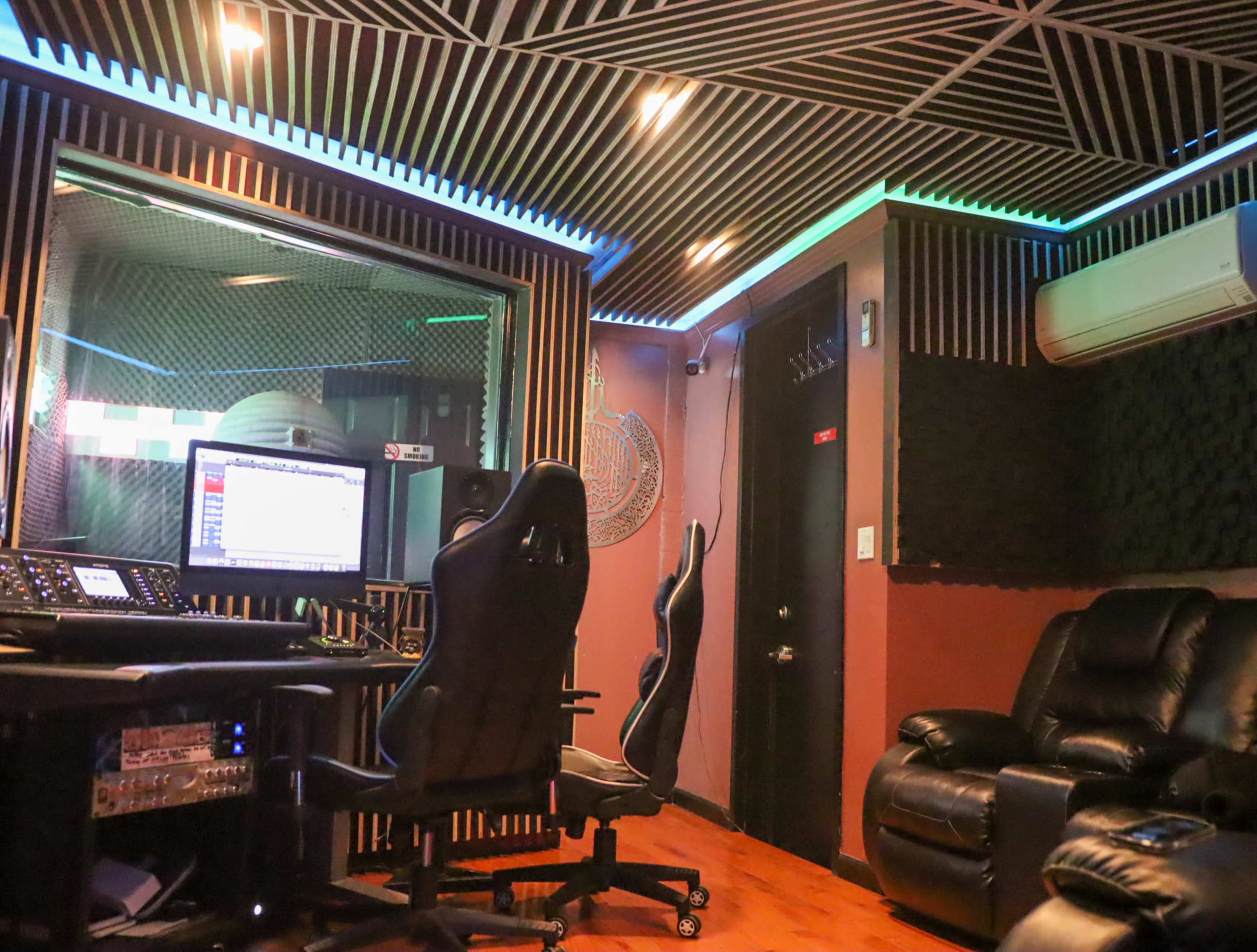 The image shows a recording studio with soundproofing, a control desk, two office chairs, and a dark leather recliner.