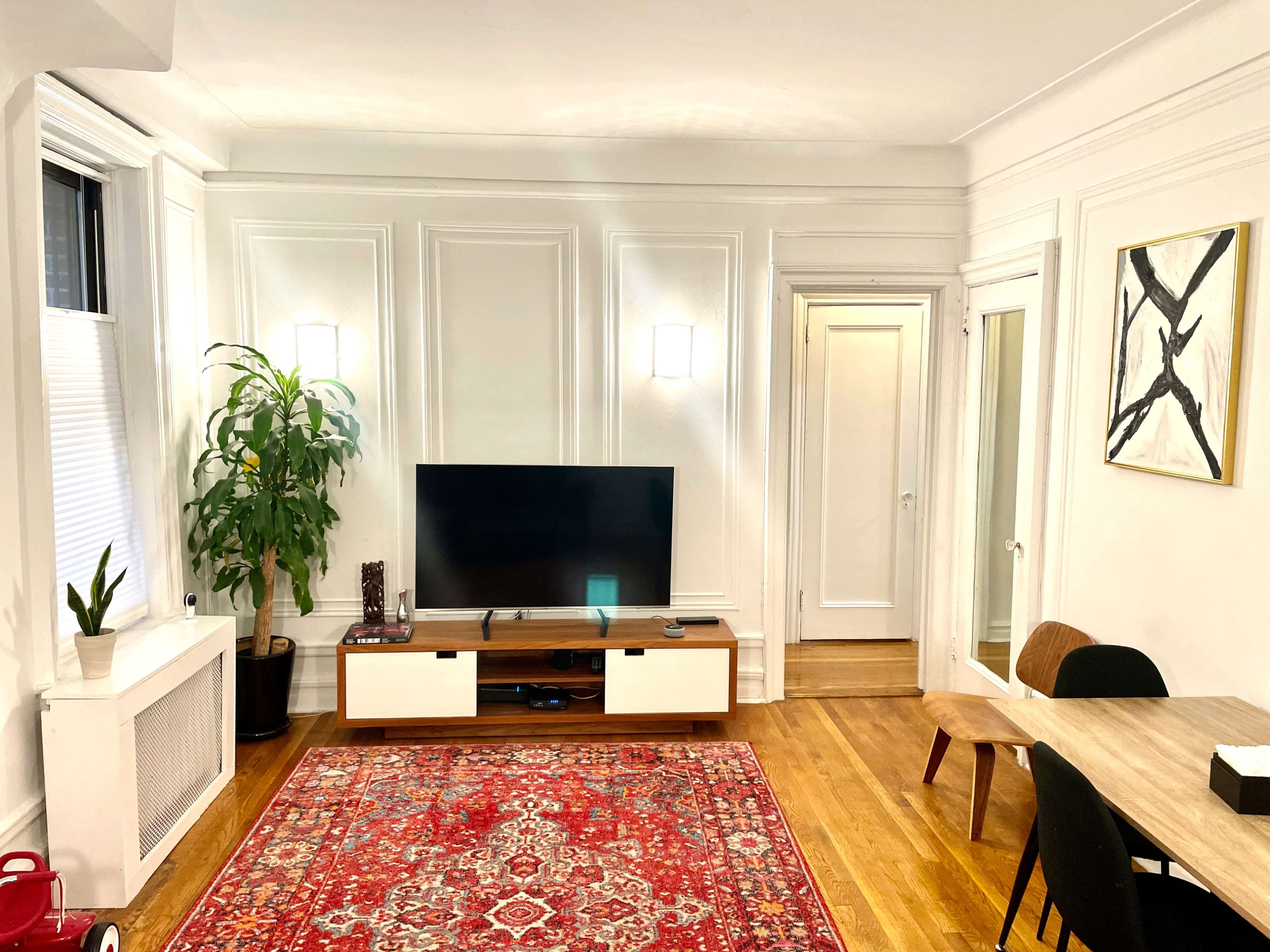 A living room with a television on a white cabinet, a large plant in the corner, a decorative rug on the wooden floor, and a dining table adjacent to a doorway.