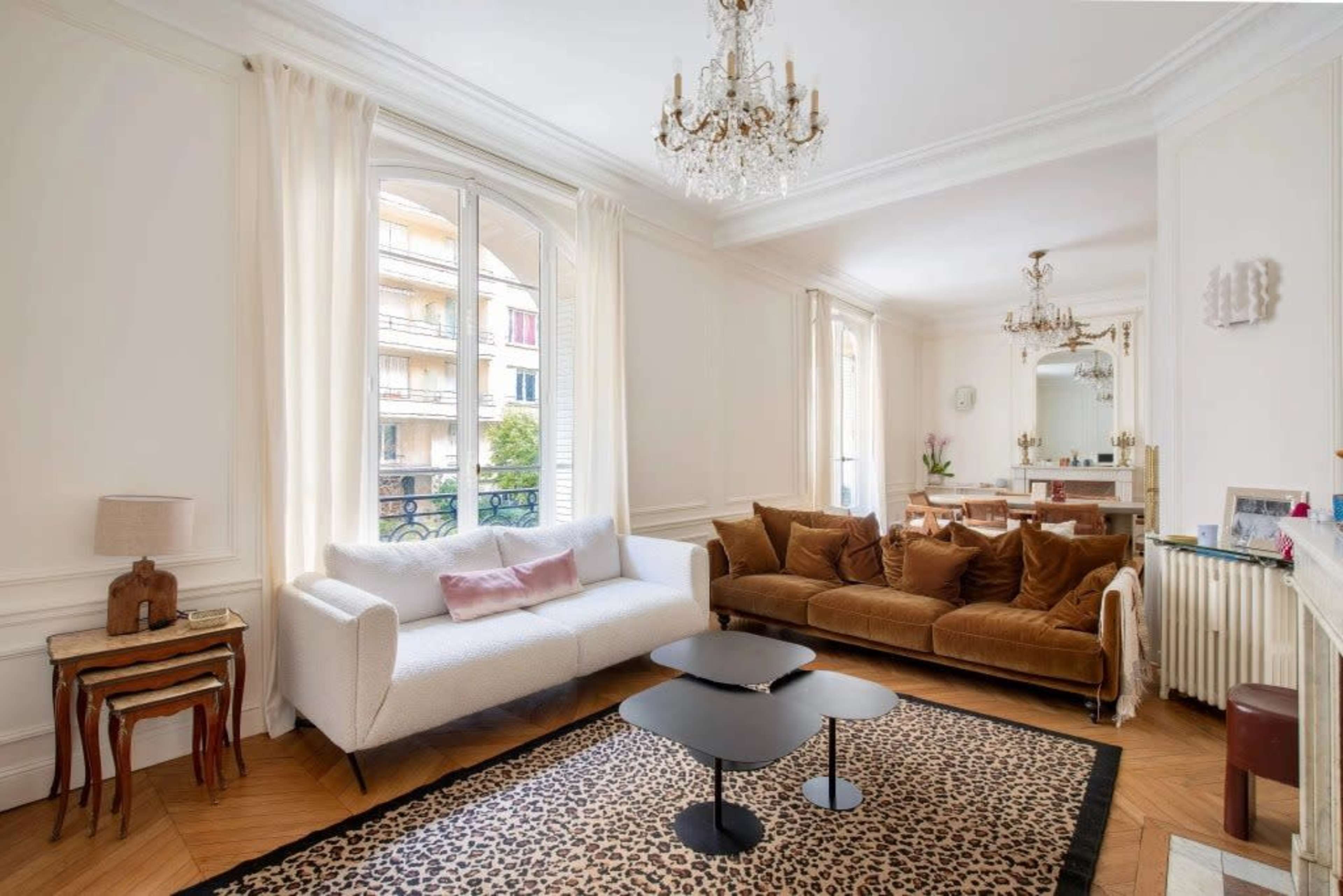 The image shows a brightly lit living room featuring a white sofa, a brown velvet couch, a black coffee table, a leopard print rug, and a dining area in the background.