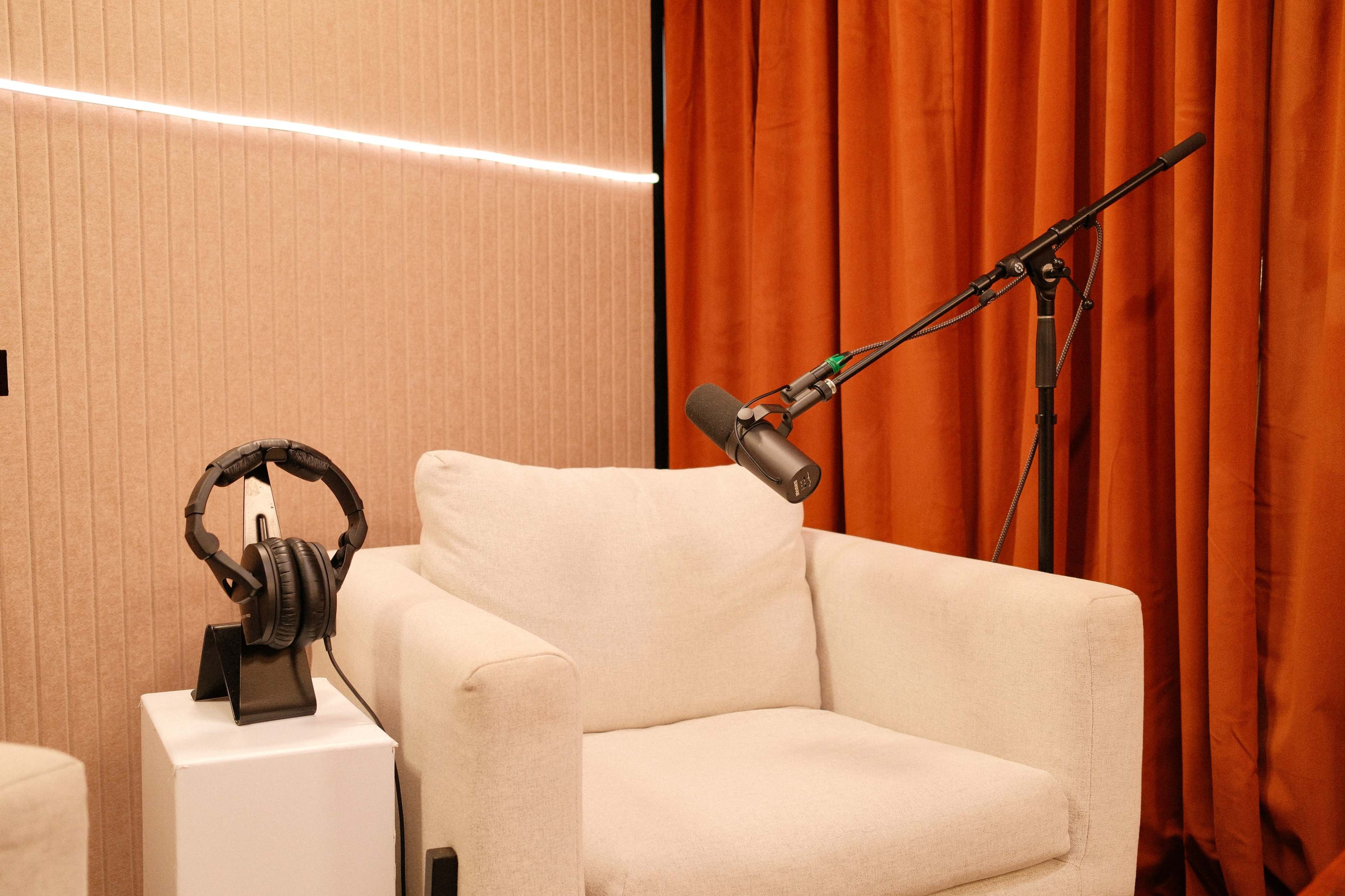 The image shows a white armchair next to a stand with headphones, in a setting with orange curtains and a microphone on a stand.