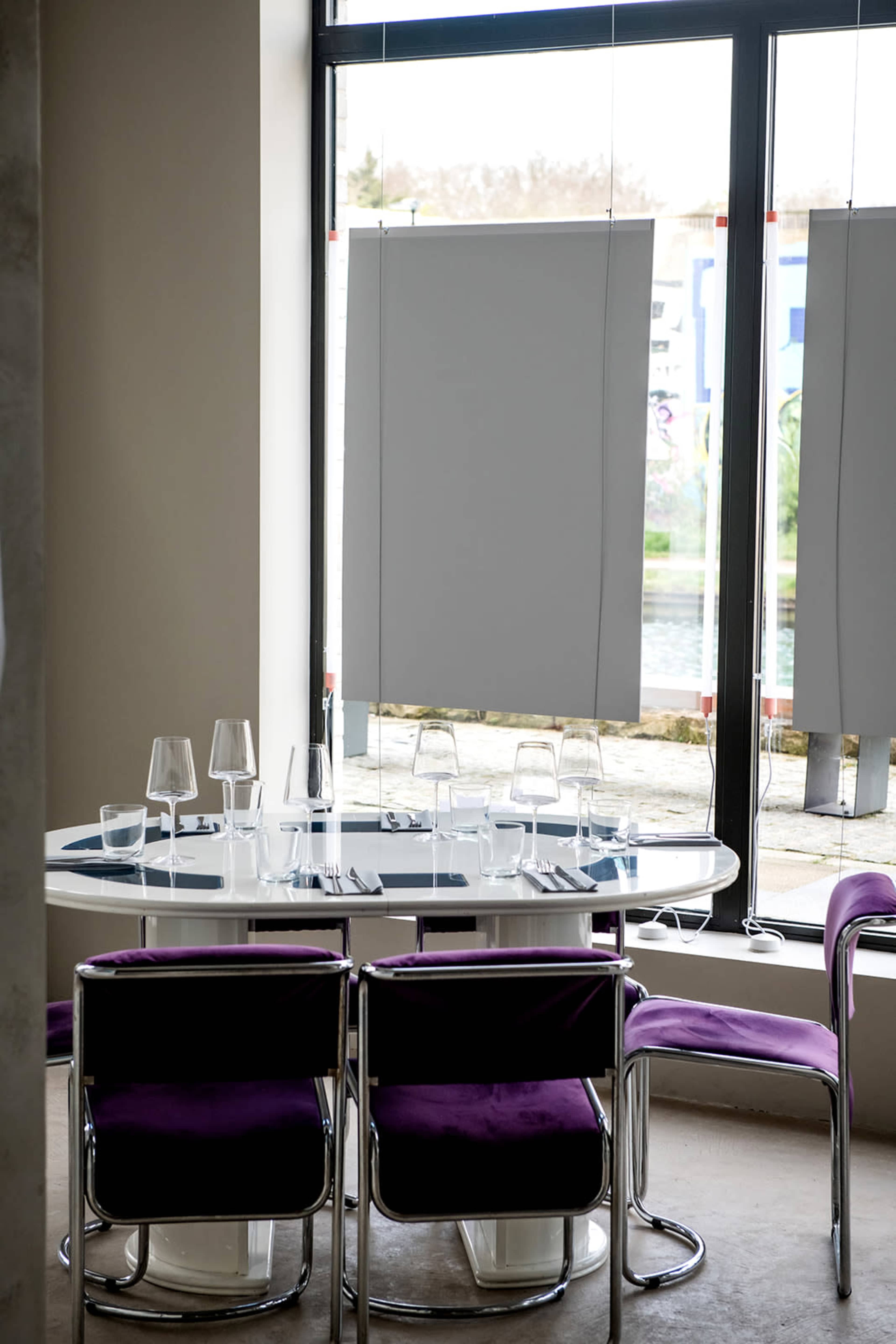 A modern dining setup features a round table with glassware and five purple chairs near a large window with partially drawn shades.