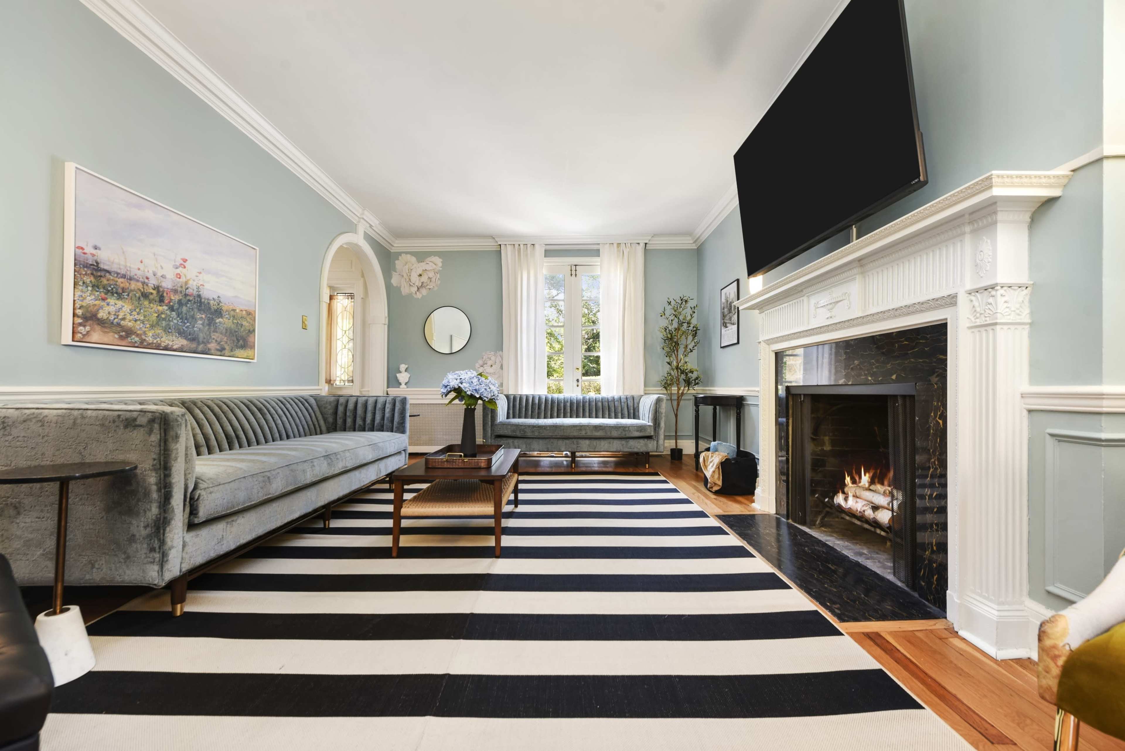 The living room features a light blue wall, two grey sofas, a striped black and white rug, and a fireplace with a flat-screen TV above it.